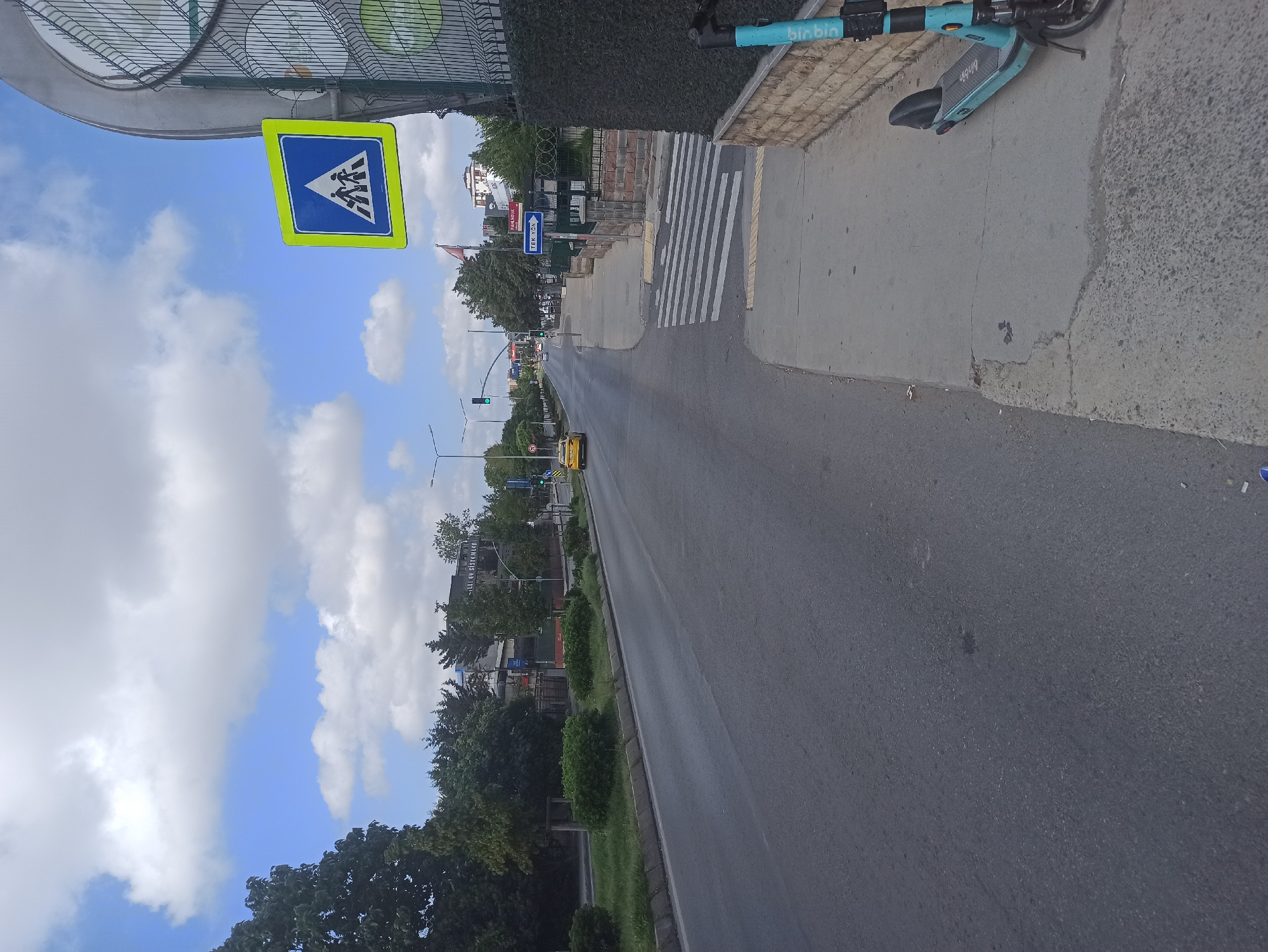 This image depicts a street scene in Ümraniye, Turkey, likely on a cloudy day. A multi-lane asphalt road stretches into the distance, with a yellow taxi visible in the left lane. The right side of the road features a sidewalk with a pedestrian crossing marked by white stripes. A bright blue electric scooter is parked on the sidewalk. Behind the sidewalk, a low stone wall topped with a dark hedge runs along the property line. In the background, trees line the street, and buildings are visible, one of which has a sign that appears to read "ALLİ GÜVEN SİSTEMLERİ." A yellow pedestrian crossing sign with a blue border is mounted on a pole to the right, overlooking the road. The sky above is a mix of blue and white clouds.