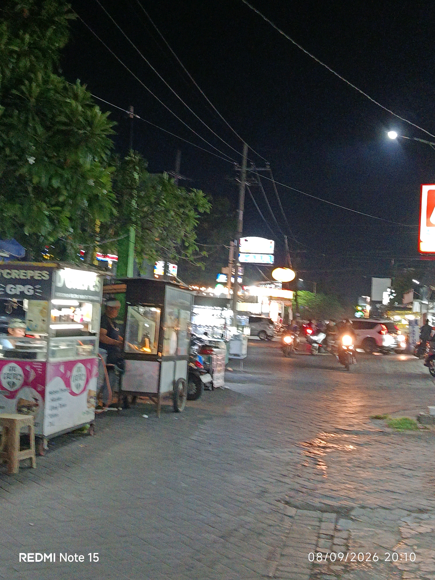 The image captures a night scene on a street in Keboansikep, Indonesia, likely a commercial area. Several food stalls are set up along the roadside, with one stall in the foreground advertising "CREPES" with colorful signage. A wooden stool is placed near this stall. Adjacent to it is another stall with a glass display case, possibly selling food items. A person is visible behind the counter of the "CREPES" stall, wearing a cap. Another person is tending to the adjacent stall. The street itself is paved with cobblestones and appears wet in places, reflecting the streetlights and vehicle headlights. Several motorcycles and some cars are present on the street, with some motorcycles in motion, their headlights illuminated. Overhead power lines crisscross the dark night sky, and a streetlamp is visible on the right side of the image. The overall atmosphere suggests a bustling street food scene at night.