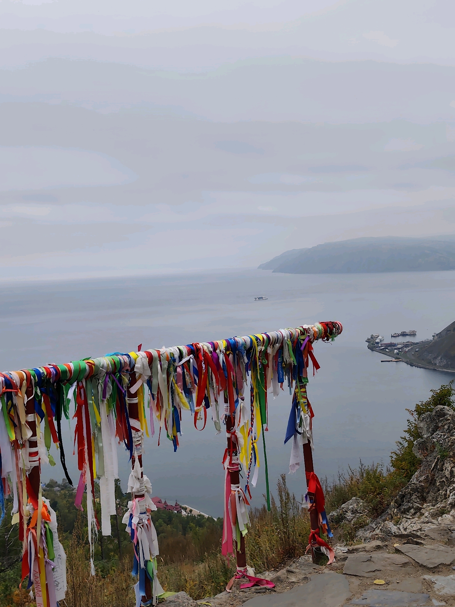 In Nikola, Russia, a scenic overlook features a railing adorned with numerous colorful prayer ribbons. The ribbons, made of fabric, are tied in bunches and hang down, creating a vibrant display against the muted tones of the sky and water. The railing itself is a metal pole, likely part of a viewing platform.
Beyond the railing, a vast body of water stretches to the horizon, with distant hills visible on the far shore. The water is a calm, steely gray, reflecting the overcast sky. Several boats are scattered on the water, including a large white vessel in the mid-ground and a cluster of ships docked at a port to the right.
The immediate foreground consists of a rocky, uneven terrain with dry grass and shrubs. The overall atmosphere is one of natural beauty and contemplative stillness, enhanced by the spiritual element of the prayer ribbons.