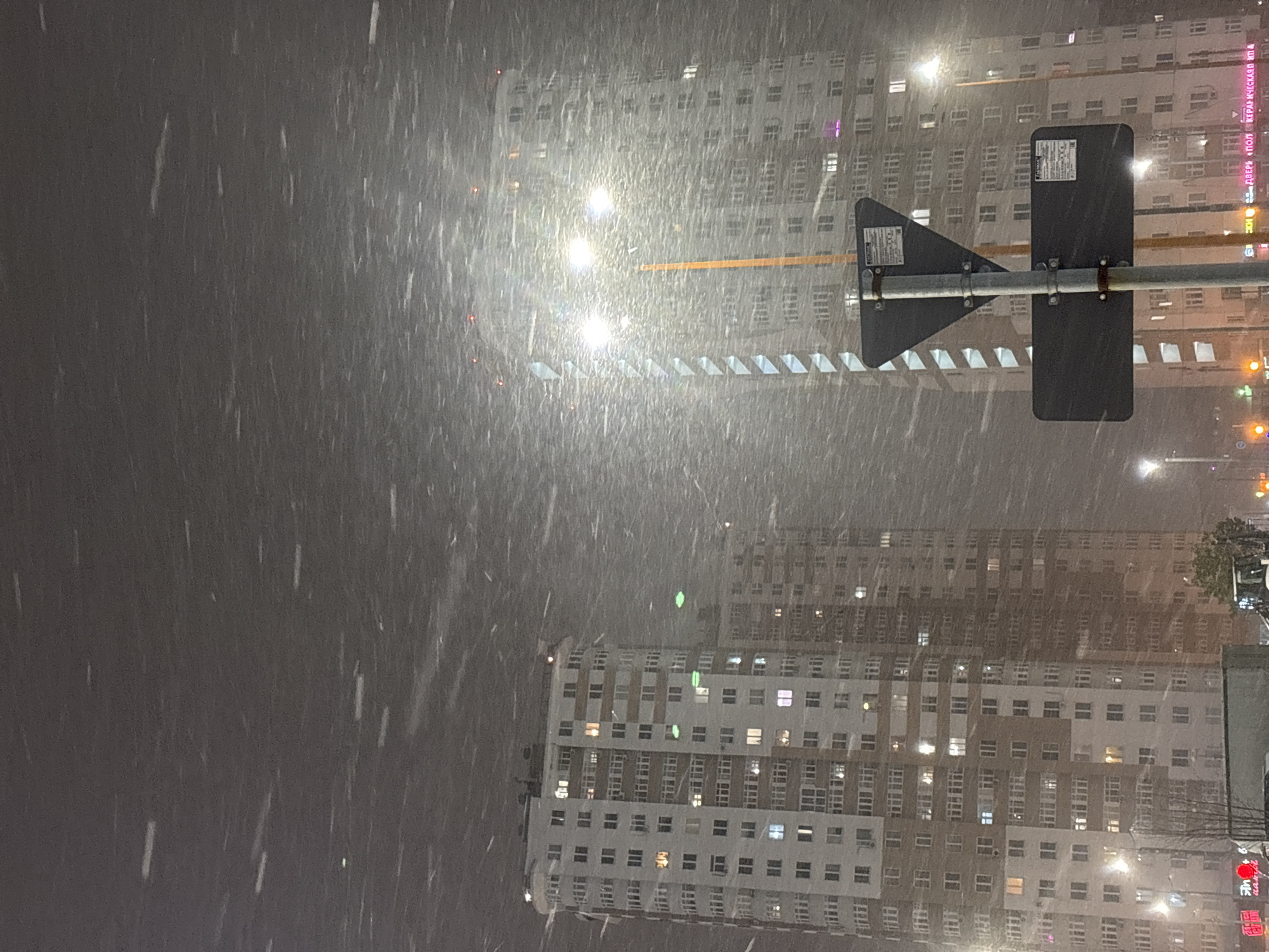 The image depicts a snowy night scene in Perm, Russia. Focus is on the road sign. Behind, tall buildings with numerous illuminated windows are visible. The heavy snowfall obscures the background details. A sign indicating a pedestrian crossing is in the foreground. Traffic lights are visible on the right side of the buildings. Overall, the image conveys a wintry urban atmosphere.