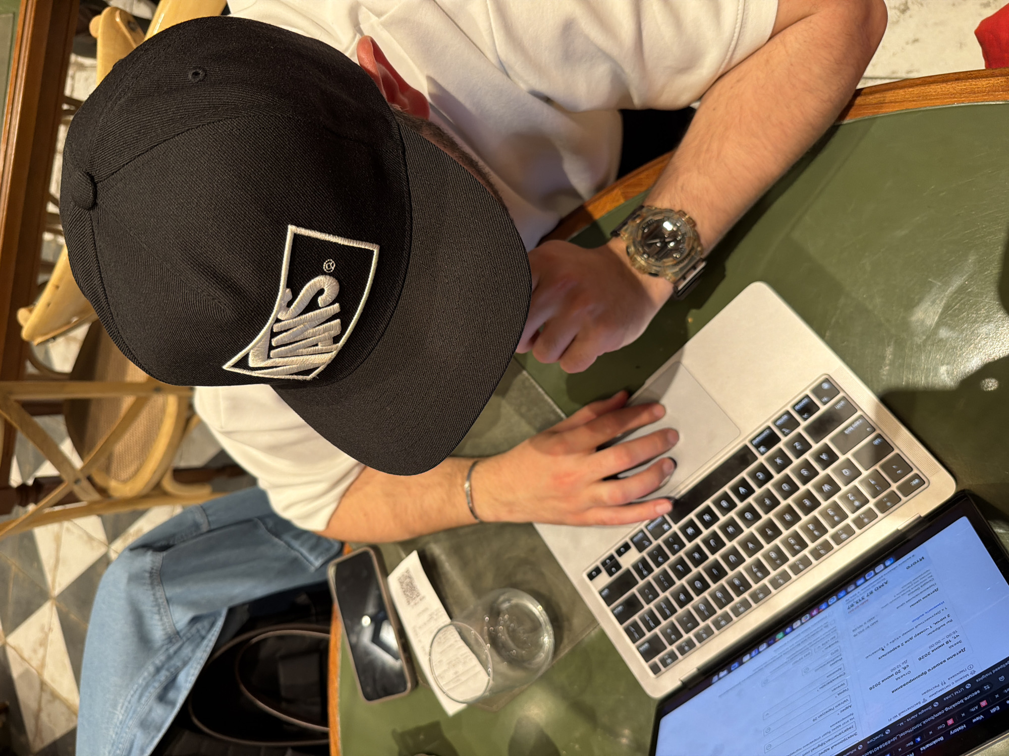 Here is a description of the image:
An overhead shot shows a person seated at a table, engrossed in their laptop. The individual wears a black baseball cap with a white logo, a white T-shirt, and a watch on their left wrist. They have a silver bracelet on their left wrist. The person's hands are positioned on the laptop's keyboard and trackpad. The laptop screen displays what appears to be a booking form or webpage in Russian, with fields for personal information. To the left of the laptop is a cell phone, a receipt, and a glass of water. The table is green, with other objects placed on it. In the background are a portion of a chair with a woven back and seat, and blue jeans. The laptop screen shows that the location is the city of Yerevan, Armenia, specifically Vahram Papazyan 29.