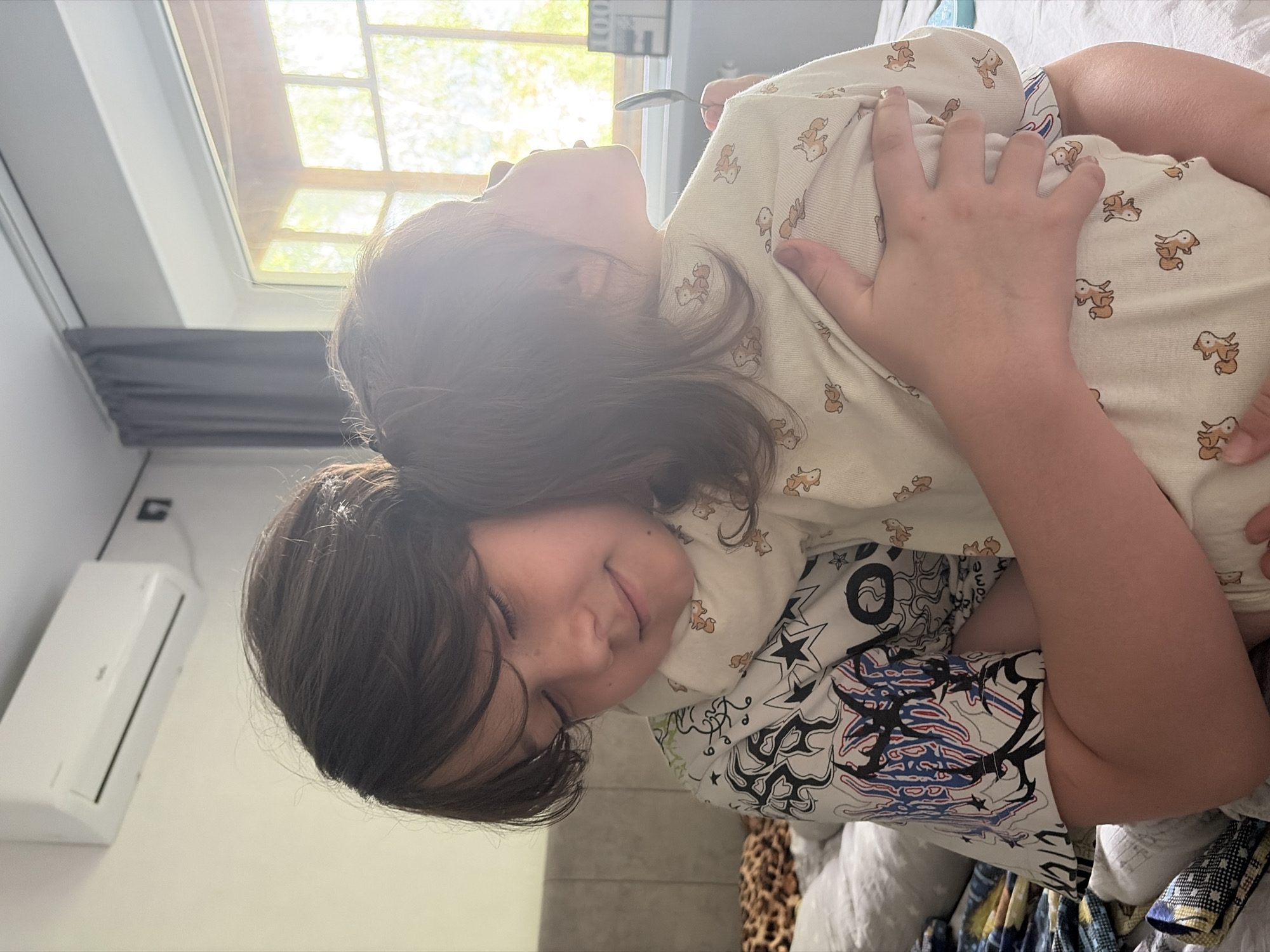 An outdoor view from inside a room shows two children being embraced by an older child. One child, seen from the back, is wearing a pajama top with a repeated pattern of cartoon foxes. They are being held by an older child with dark hair and closed eyes. This older child is wearing a white top with a graphic design and a white scarf with a fox pattern. The background includes a white air conditioner mounted on the wall, a grey curtain, and a window that reveals a blurry green outdoor scene. The children are on a bed covered with white and patterned bedding. The overall scene suggests a domestic setting, possibly a bedroom, with natural light streaming in. The location is identified as Chaikovsky, Russia.
