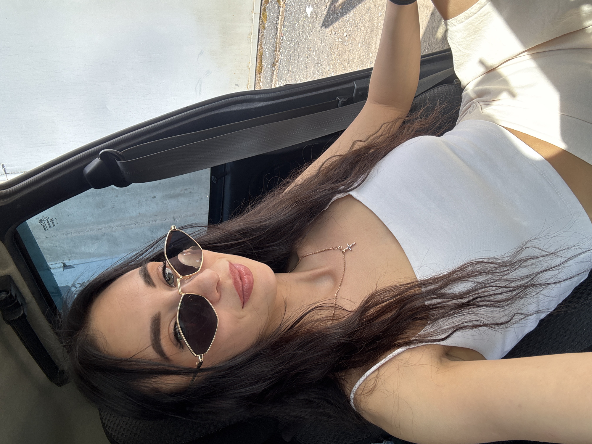 A young woman is lying in a convertible car, presumably in Chaikovsky, Russia, based on the prompt. She is wearing sunglasses, a white top, and light-colored shorts. The car is parked, and the background shows a combination of a building exterior and pavement. She wears a necklace with a cross pendant. The lighting suggests it is daytime.