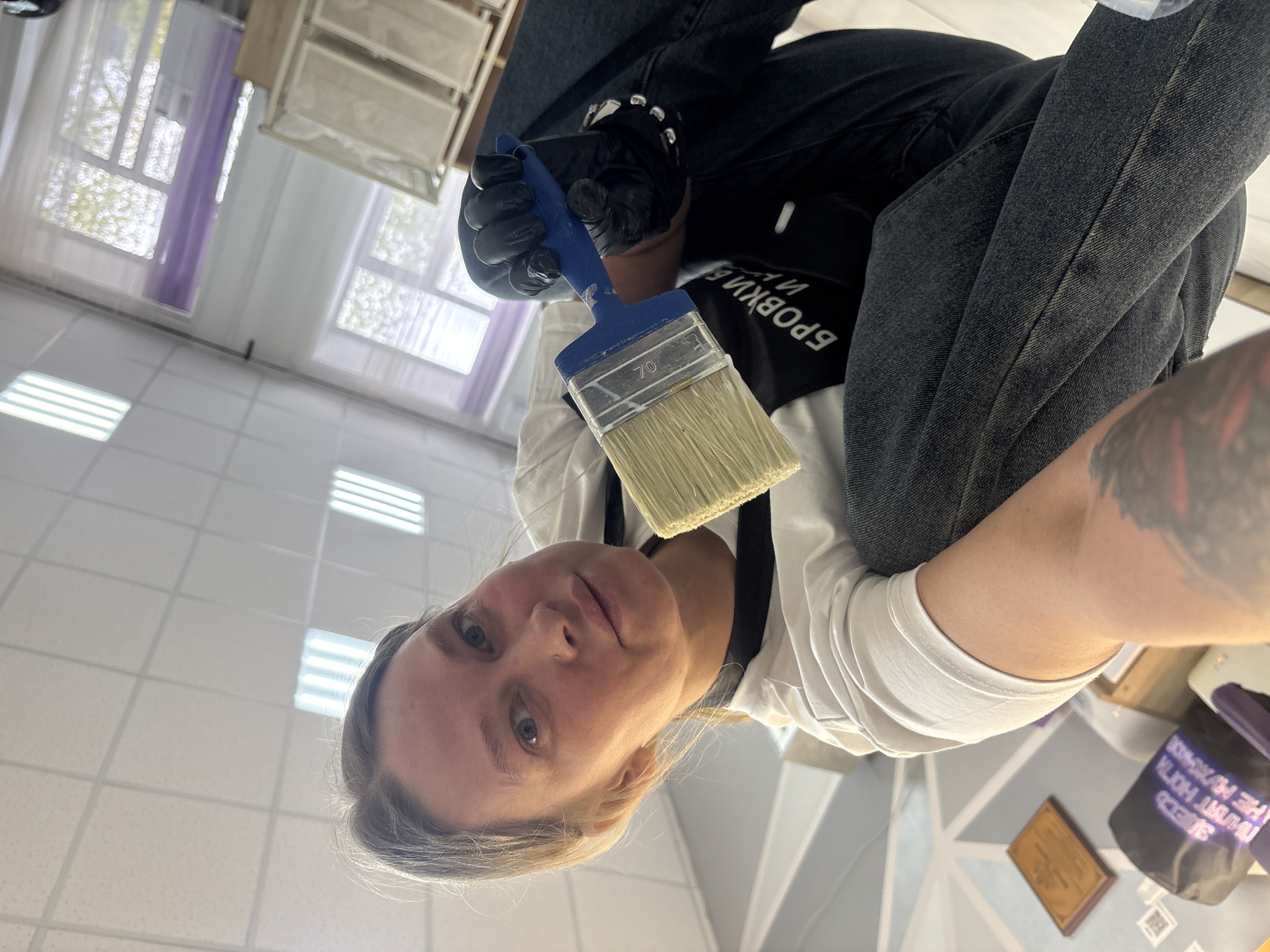 A person, likely female, is holding a paint brush in a black-gloved hand, angled towards the camera. She has fair skin, light hair, and blue eyes. She is wearing a white shirt and a black apron with text in Russian, likely related to eyebrows. She also wears dark jeans. The paint brush has a blue handle and natural bristles. The scene is indoors, with tiled flooring and ceiling with fluorescent lights. Behind the person, there is a window with white curtains. The setting appears to be a beauty salon. The location is Perm, Russia, as indicated by the Russian text on the apron. There is a table behind with some objects on it, possibly containers for beauty products.
