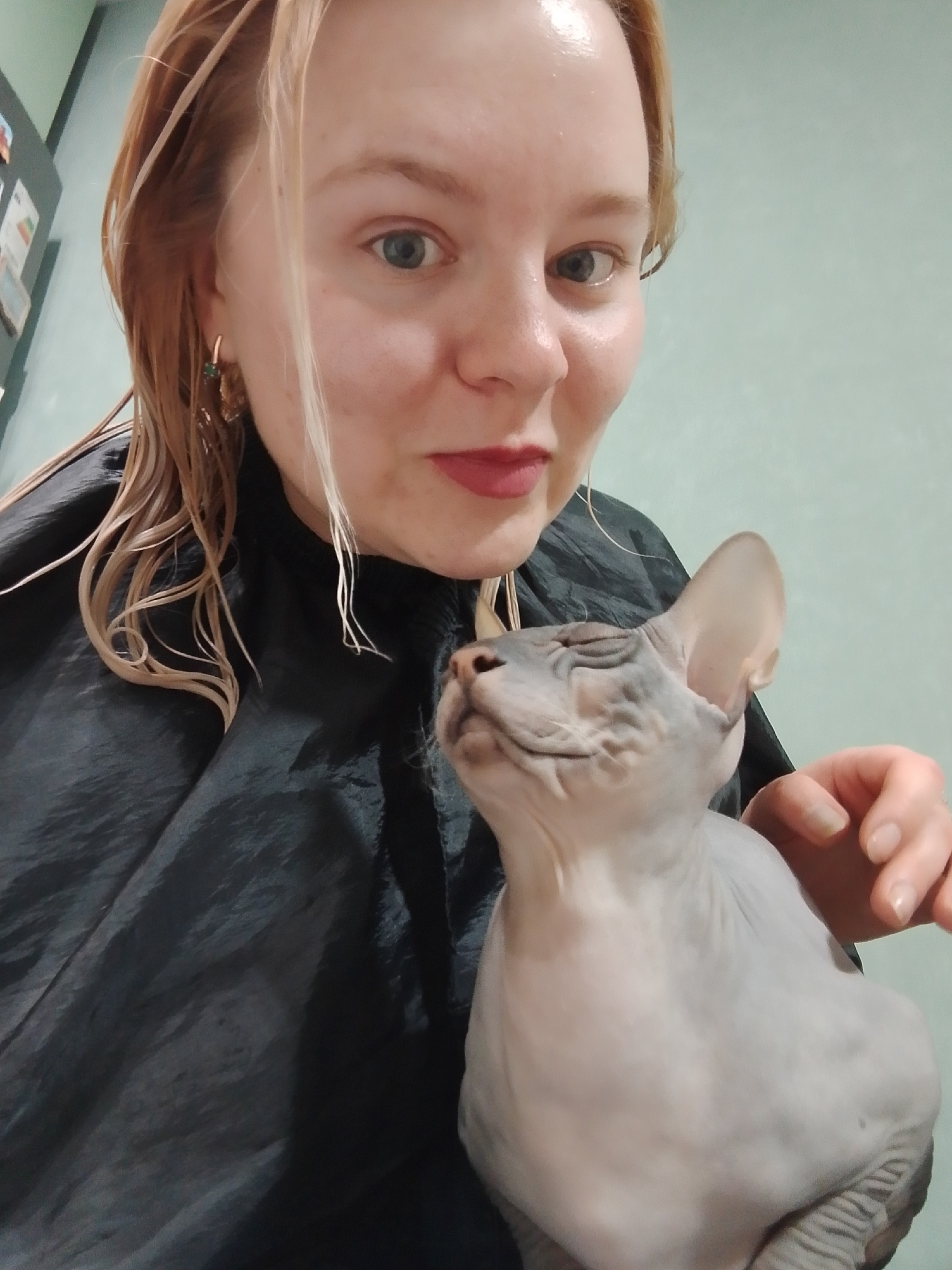 A fair-skinned woman with wet blonde hair and blue eyes is holding a sphynx cat. She wears red lipstick and earrings, and is draped in a black material. The cat is looking upwards with its eyes nearly closed. The background is a light green wall. The location is unknown, the city of Izhevsk, Russia cannot be confirmed.