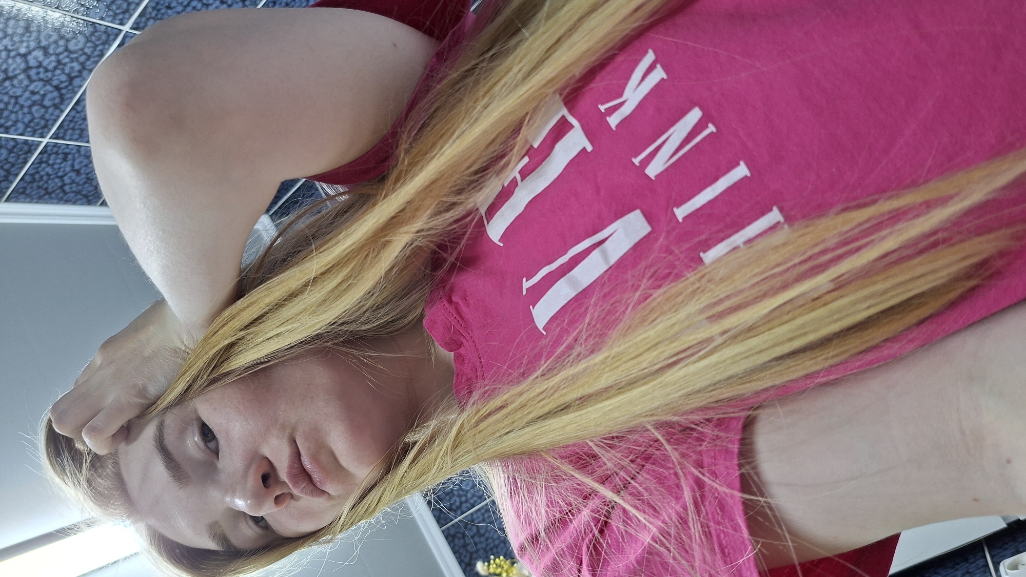 A woman with long blonde hair is lying down, wearing a pink t-shirt with the word "PINK" printed on it in white lettering. She is holding her hand near her forehead. The background shows a bathroom with blue and white tiled walls and a glimpse of a yellow flower arrangement. The image was taken in the city of Chaikovsky, Russia.