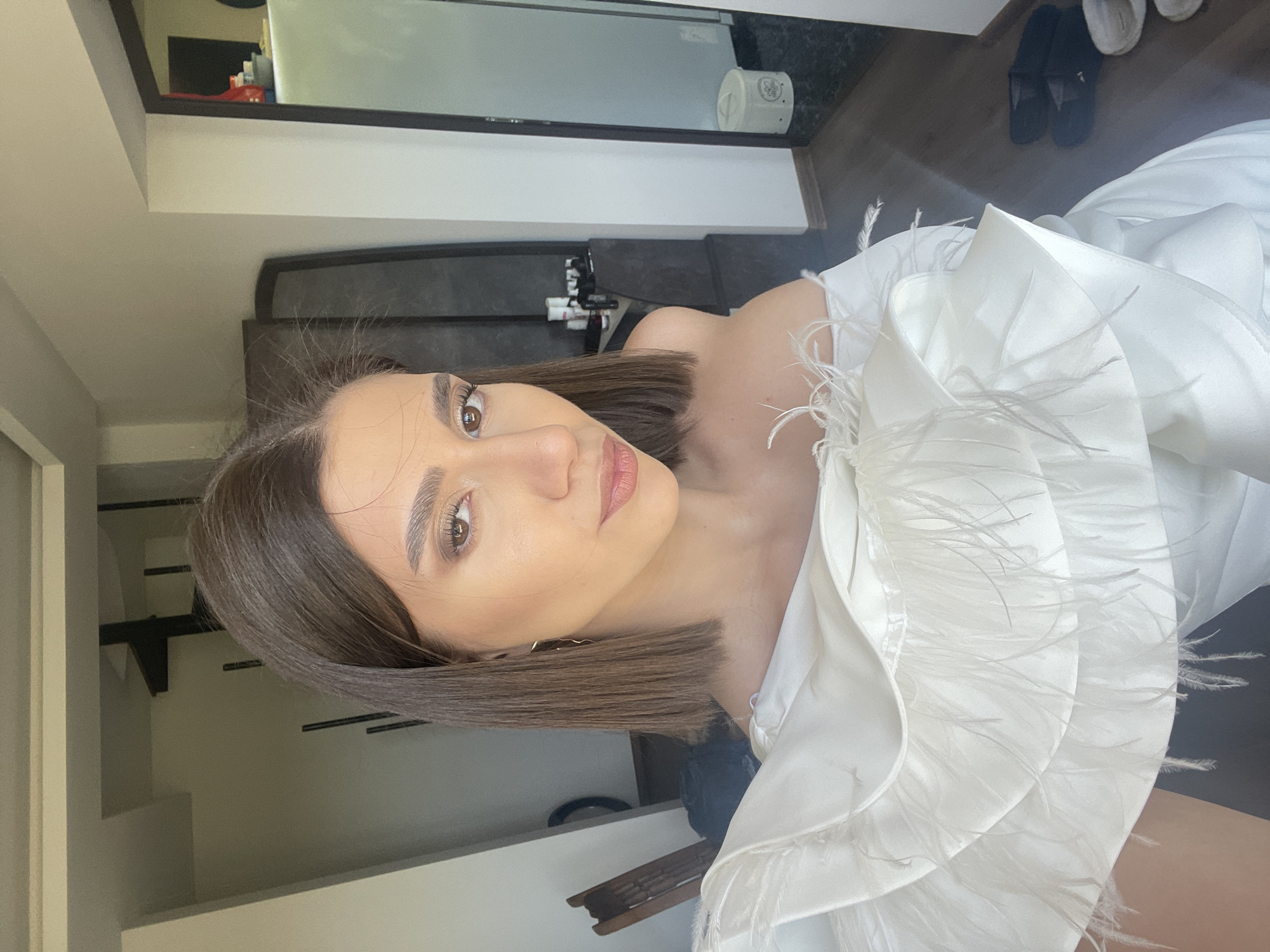 The image depicts a woman with brown, shoulder-length hair, wearing a white dress adorned with feathered details around the neckline. She appears to be indoors, possibly in a residential bathroom, given the presence of a mirror, vanity, and bathroom accessories. Slippers are seen on the floor in the background. It is not possible to determine if the picture was taken in Sofia, Bulgaria.