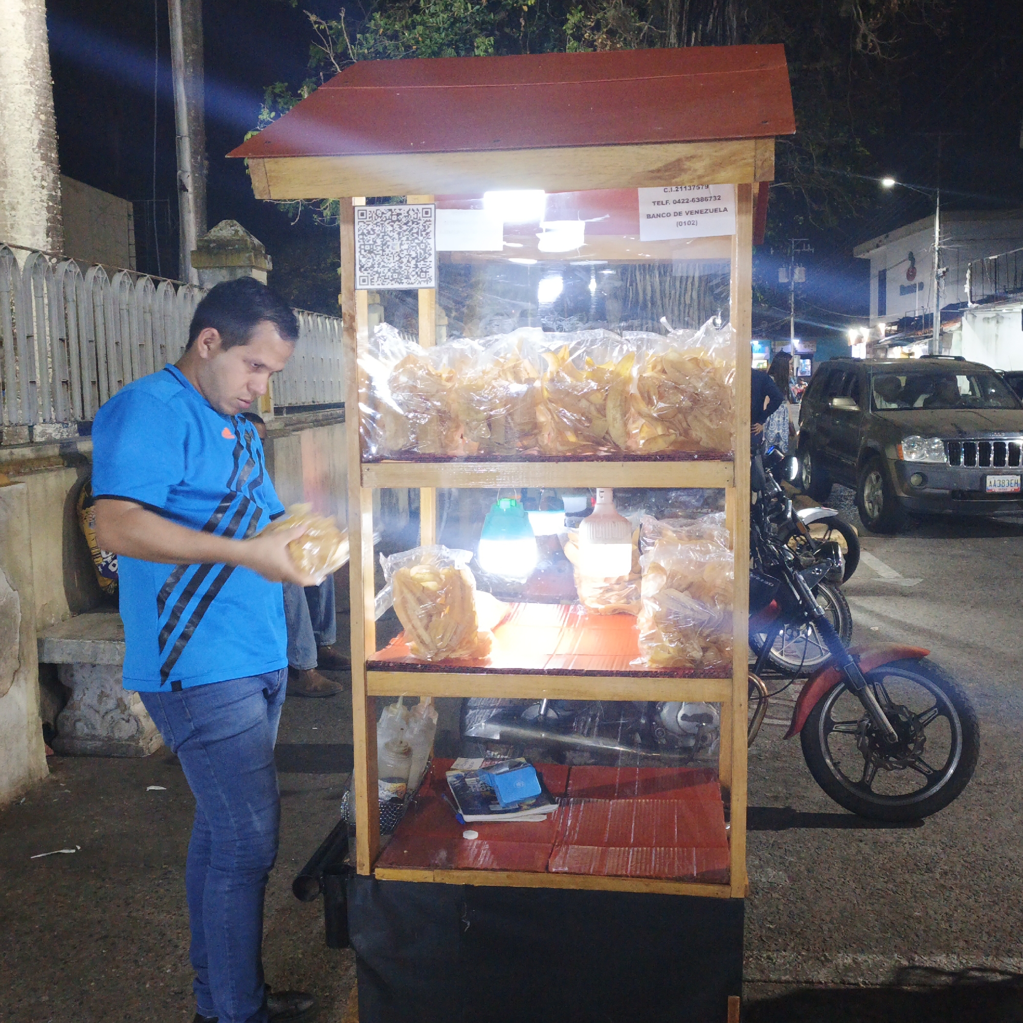 At night in Tinaquillo, Venezuela, a man in a blue shirt and jeans stands next to a wooden food stand on the street. The stand is stocked with bags of chips, illuminated by a light on the middle shelf. The stand also has a QR code and banking information displayed. Behind the stand, there is a motorcycle and a dark green SUV. Other people and buildings are visible in the background, along with streetlights. The car license plate reads AA383EH.