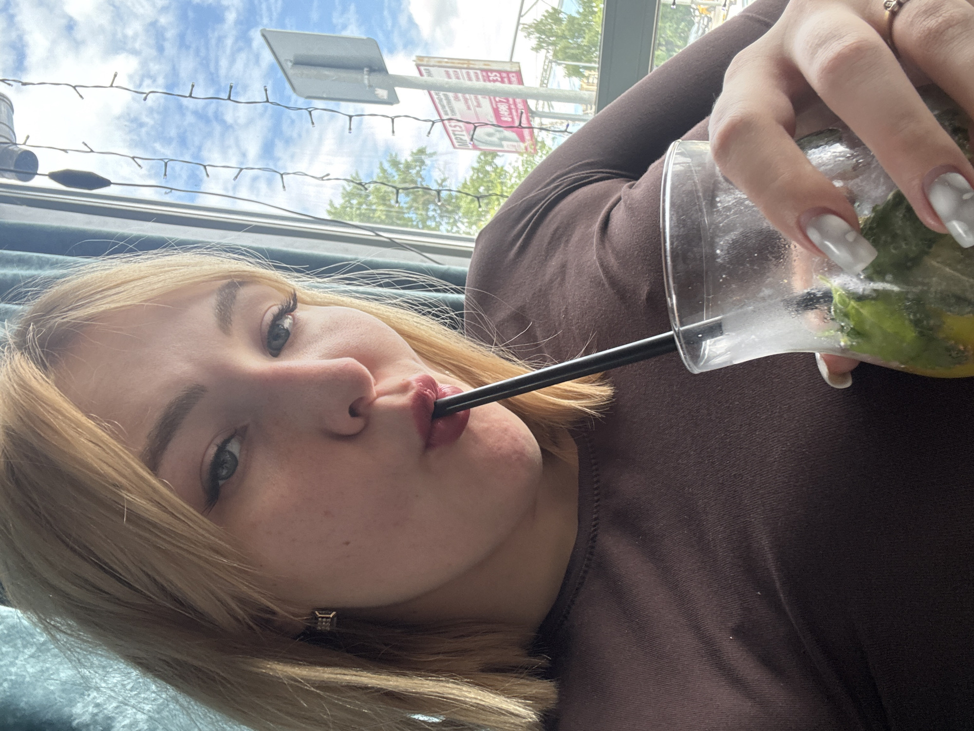 A young woman with blonde hair is captured from the chest up, sitting indoors near a window. She is holding a glass filled with a green beverage and mint leaves, drinking through a black straw. She wears a brown long-sleeved shirt. Her facial expression is relaxed as she sips her drink. Through the window, a bright sky with clouds is visible, along with trees and a sign that reads "MPTLS" in Russian. The scene appears to be in an establishment in Khimki, Russia, possibly a cafe or restaurant, given the indoor setting and view outside.