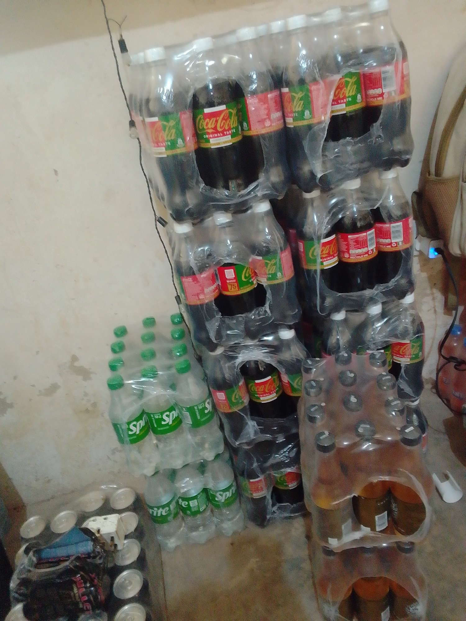 The image depicts a storage area, possibly a corner of a room or a small shop, with several stacks of bottled beverages. The primary focus is on a large collection of Coca-Cola bottles, primarily in their standard red and green packaging. These bottles are shrink-wrapped in plastic, forming multiple columns and rows, suggesting they are intended for sale or distribution.
Adjacent to the Coca-Cola stacks, there are packs of Sprite bottles, identifiable by their green caps and labels. These are also shrink-wrapped. Further to the right, there is a stack of what appear to be Fanta bottles, with orange-colored liquid visible through the clear plastic bottles and packaging.
In the foreground, to the left, a collection of cans is visible, partially covered by a plastic wrap and a dark fabric. There is also a small white object and other miscellaneous items.
The environment appears to be an indoor space with a neutral-colored, textured wall in the background. The floor is also visible, appearing to be concrete or a similar material. The lighting is somewhat dim, casting shadows and highlighting the plastic packaging of the drinks.
The presence of numerous packaged beverages suggests a commercial setting, such as a store, warehouse, or a stall in a market. Given the context provided about Jos, Nigeria, this scene is likely from a commercial establishment within the city, possibly a small business or a distributor's storage. There are no people visible in the image.