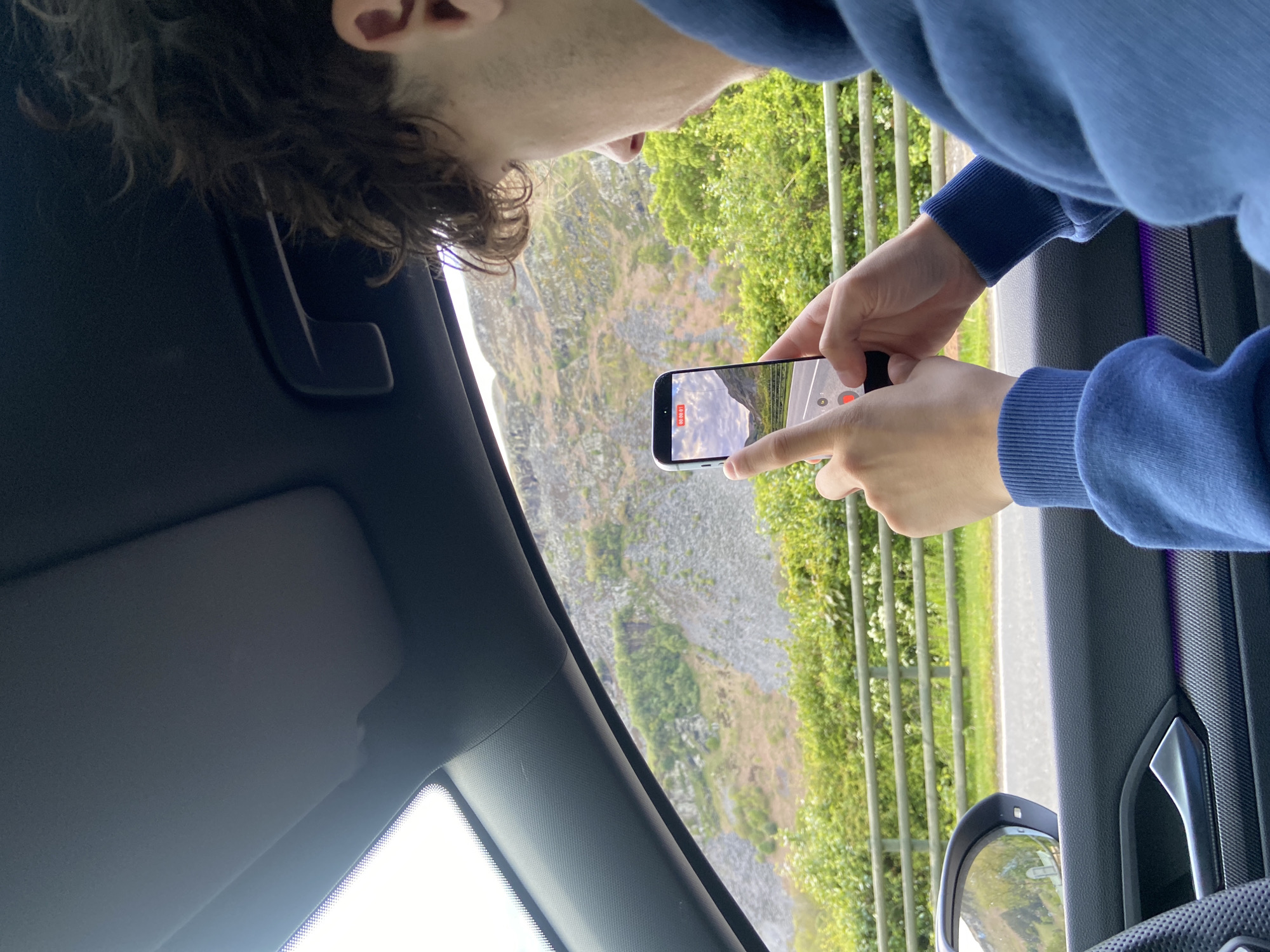 From inside a car, a person wearing a blue sweatshirt is filming a rocky hillside and a road in Llangynog, United Kingdom. The phone screen reflects the scene. The car interior is visible. The window is slightly down.