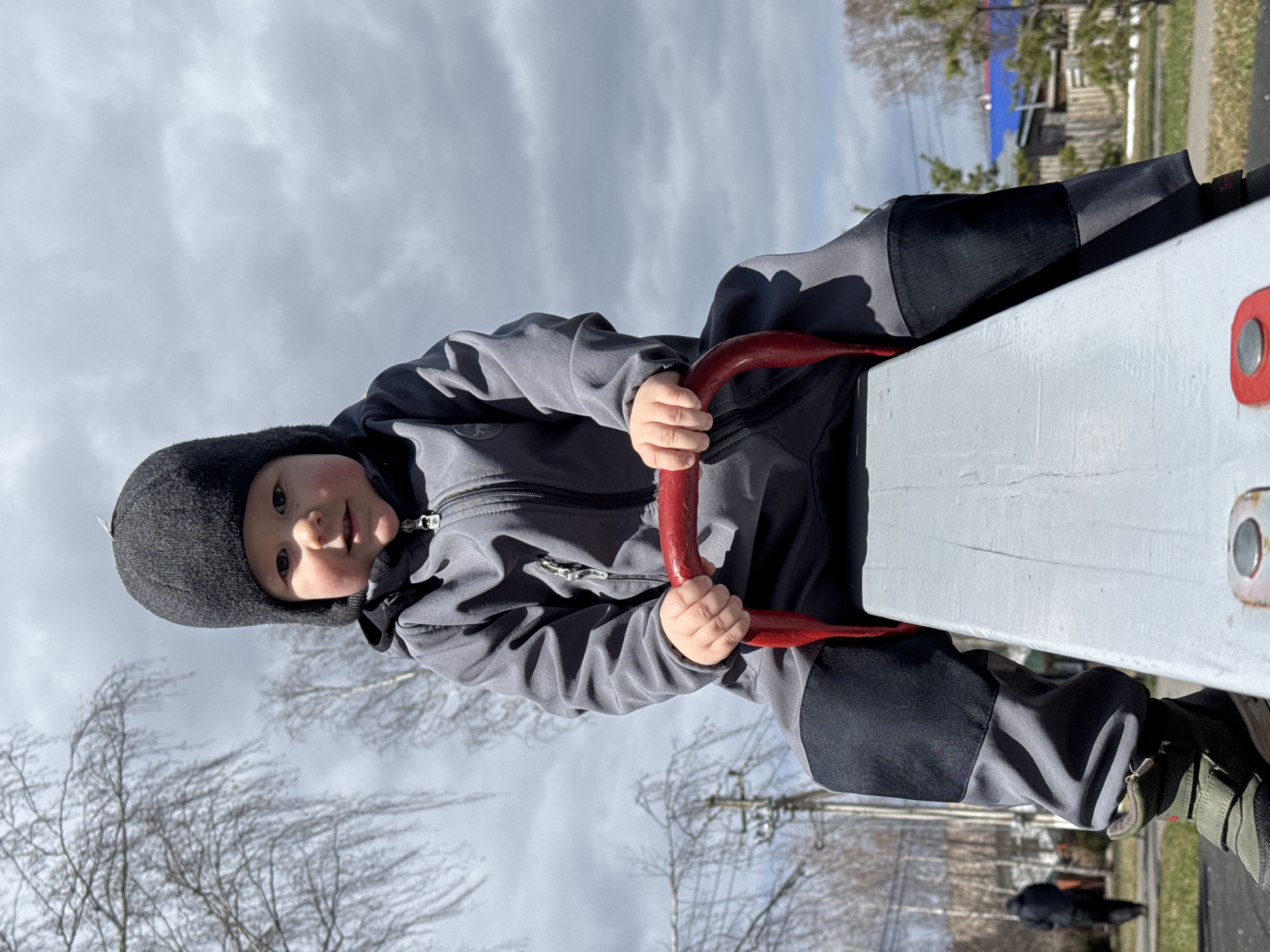 A young child, wearing a dark gray hooded jacket and a dark gray winter hat, is sitting on a white seesaw with red handles. The child's hands are gripping the red handles. The background shows a cloudy sky, bare trees, a building with a blue roof, and a grassy area. The location is potentially a park or playground. The image does not provide explicit information that the scene is in Zav'yalovo, Russia, so that part is speculative.