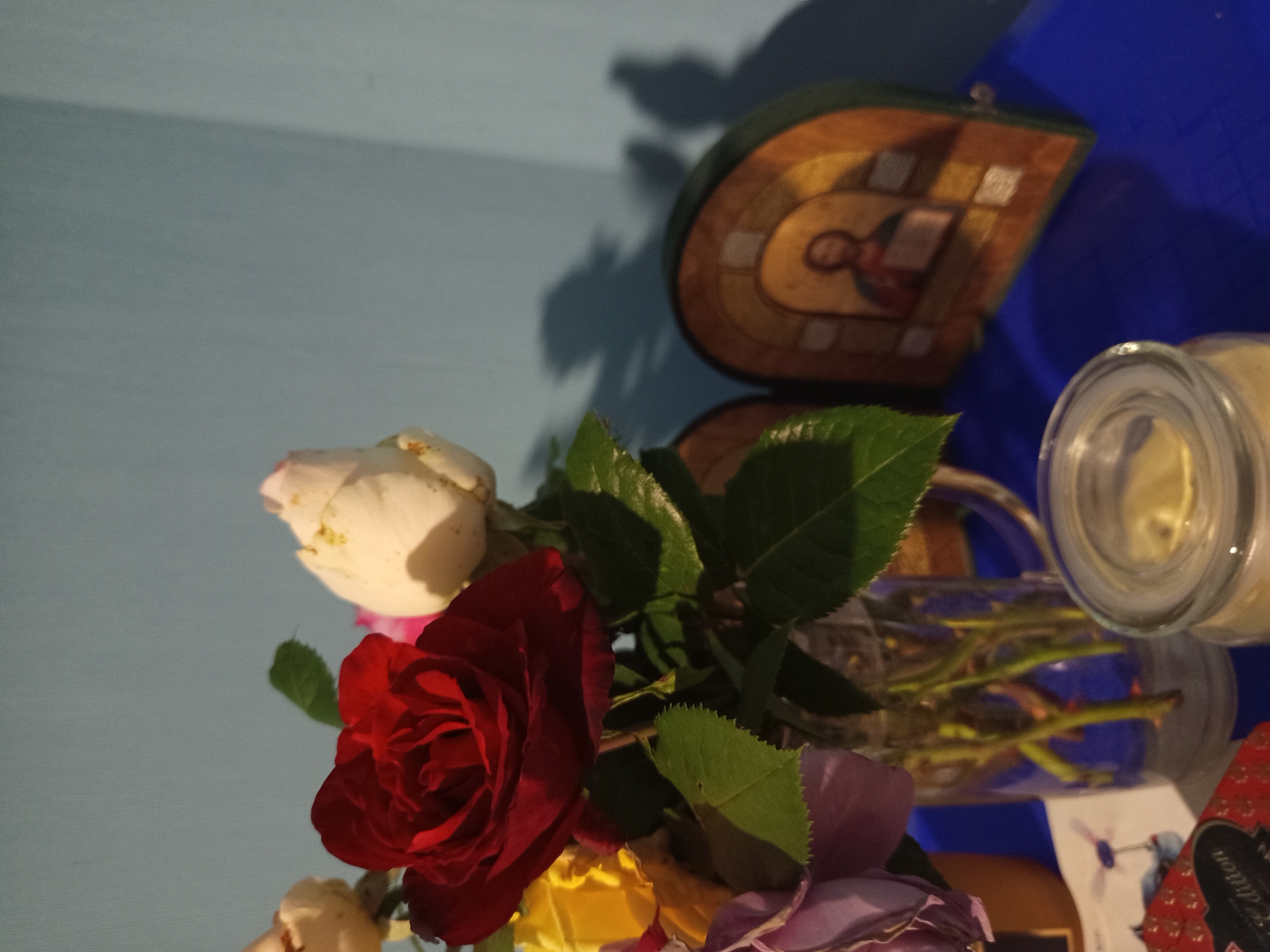 The image depicts a still life arrangement on a blue surface, likely a table or shelf, against a light blue wall. In the foreground, a clear glass vase holds a bouquet of roses in various stages of bloom and color, including red, white, yellow, and purple. Some roses are fully open, while others are still buds. Green leaves are interspersed among the flowers. To the right of the vase, a small, arched wooden icon is visible, featuring a religious depiction, likely of Jesus Christ. The icon casts a shadow on the wall behind it. In the lower right corner, a clear glass jar with a lid is partially visible. In the lower left, a red and gold patterned box and a white card with a graphic of a person riding a bicycle are also present. The lighting suggests an indoor setting, possibly with natural light casting shadows. The overall scene conveys a sense of domestic tranquility and devotional practice. There is no indication of the city of Чэрвеньскі сельскі Савет, Belarus within the image itself.