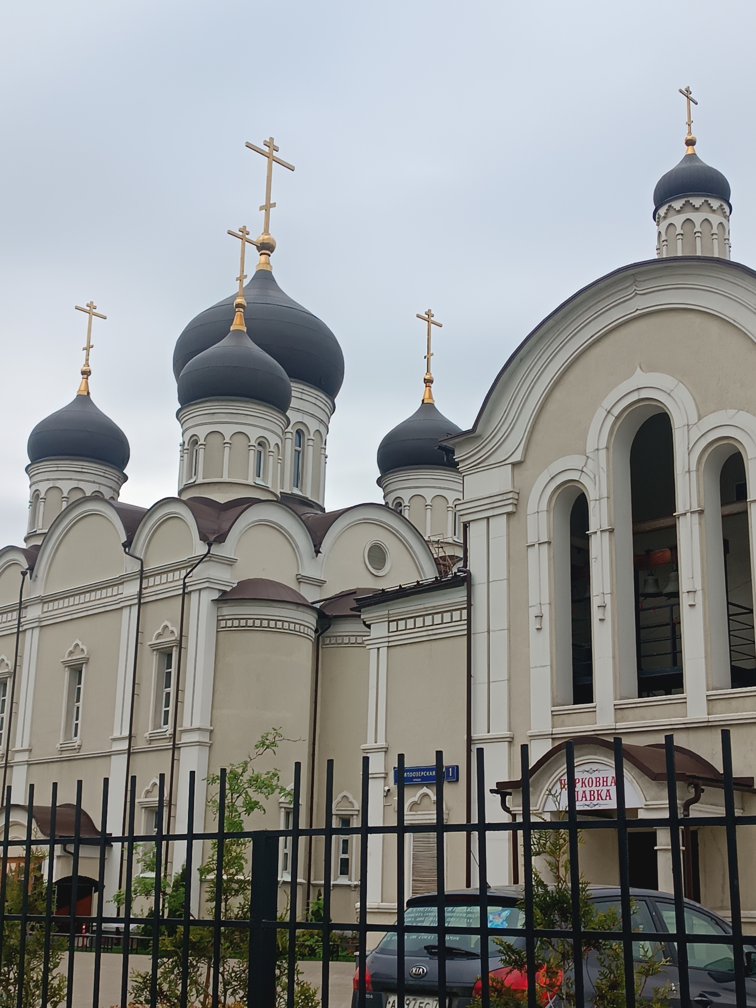 A low-angle, medium shot captures the exterior of a beige Orthodox church with black domes, adorned with gold crosses, located in Moscow, Russia. The church features multiple domes, arched windows, and decorative molding. In front of the church is a black metal fence, partially obscuring a dark-colored car. A street sign labeled "АТООЗЕРСКАЯ 1" is visible on the church wall. A sign above an entrance reads "ЦЕРКОВНАЯ ЛАВКА". The sky is overcast, providing soft, diffused light.