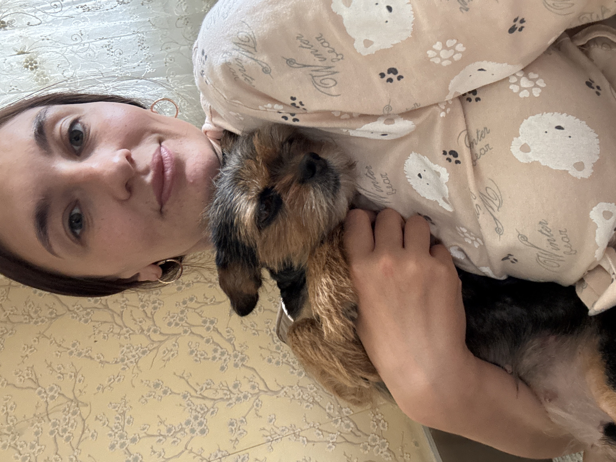 A young woman with dark hair and hoop earrings lies comfortably, smiling softly at the camera, while holding a small, brown and black wiry-haired dog against her chest. The dog is nestled closely, looking towards the camera with a gentle expression. The woman is wearing a light peach-colored top adorned with a repeating pattern of white bear heads, dark paw prints, and the cursive text "Winter Bear". The background features a light cream or yellow surface with a delicate, light-colored floral or branch pattern, likely a bedsheet or wallpaper, indicating an indoor setting. The scene captures a relaxed and affectionate moment between the woman and her pet, suggesting warmth and comfort in a home environment in Sochi, Russia.