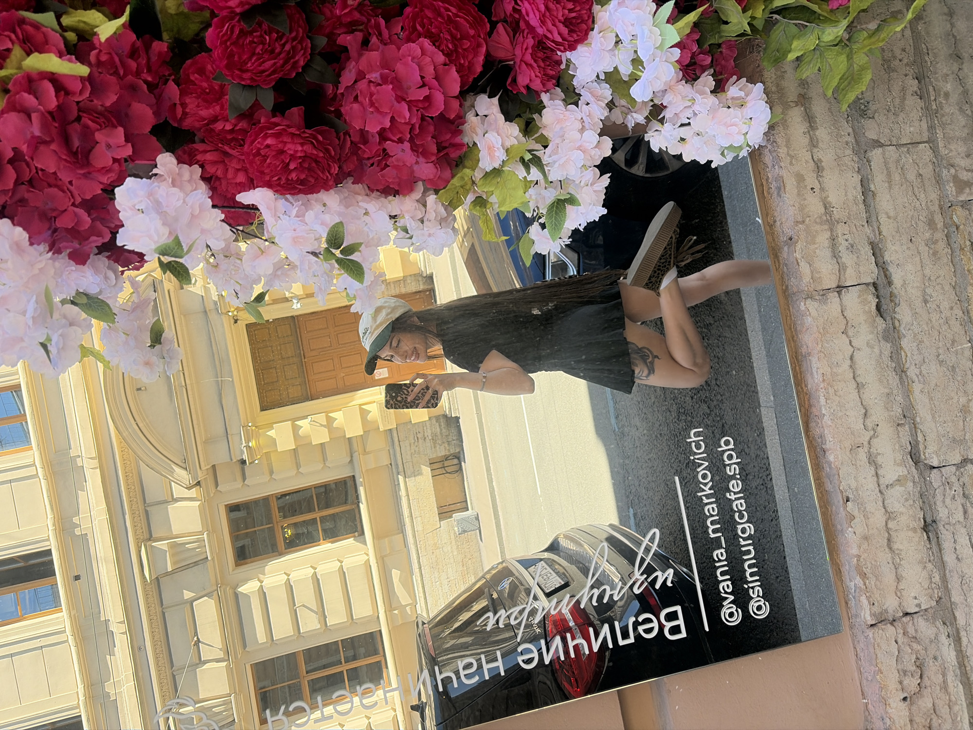 The image shows a reflection in a mirror, with a young woman in a black dress and a white baseball cap posing in front of a building. She is holding a smartphone to take a selfie. Behind her is a black car and the facade of a building with windows. The reflection also shows an arrangement of pink and white flowers. Text on a black sign in the reflection reads "@vania_markovich" and "@simurgcafe.spb". The building's architecture suggests it is located in Saint Petersburg, Russia, a city known for its historical buildings and decorative elements.