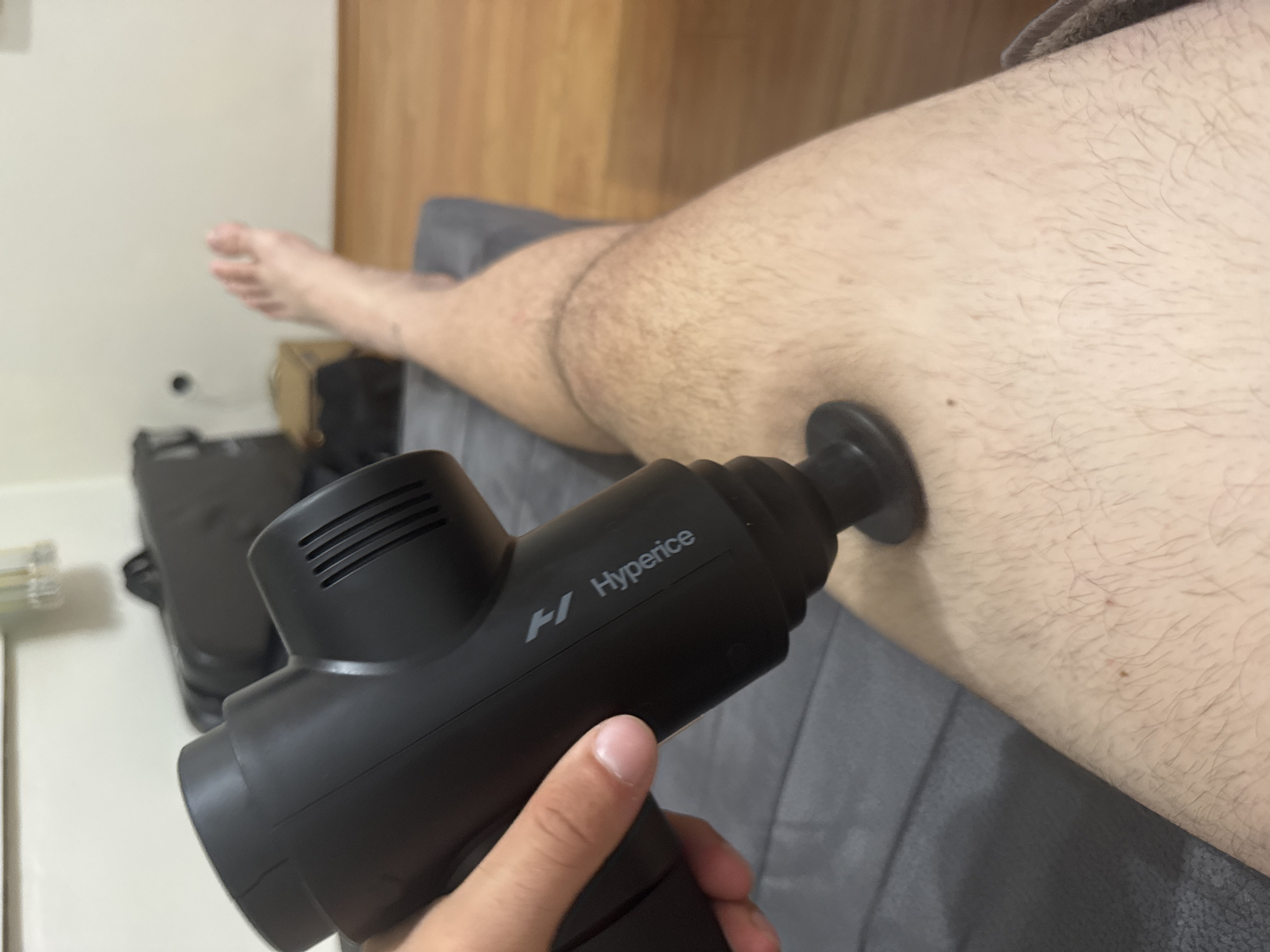 This indoor image, captured in Stara Zagora, Bulgaria, depicts a person using a massage gun on their leg. The central focus is a black "Hyperice" brand massage gun, clearly displaying its name and a stylized "H" logo. A hand with visible fingers is holding the device firmly, with its round attachment head pressed against the person's leg, suggesting active use for muscle relief or recovery.
The person's leg, covered in visible hair, is extended and resting on what appears to be a grey fabric bed or sofa. Their foot is partially visible in the upper left background. The setting is a casual indoor environment, likely a bedroom or living room. The background features a wooden wall or furniture on the right side and a plain white wall on the left, with some indistinct, blurry dark objects and a light fixture further in the distance. The lighting is ambient indoor light, providing no specific cues about the time of day. The image captures a moment of personal wellness or physical therapy.