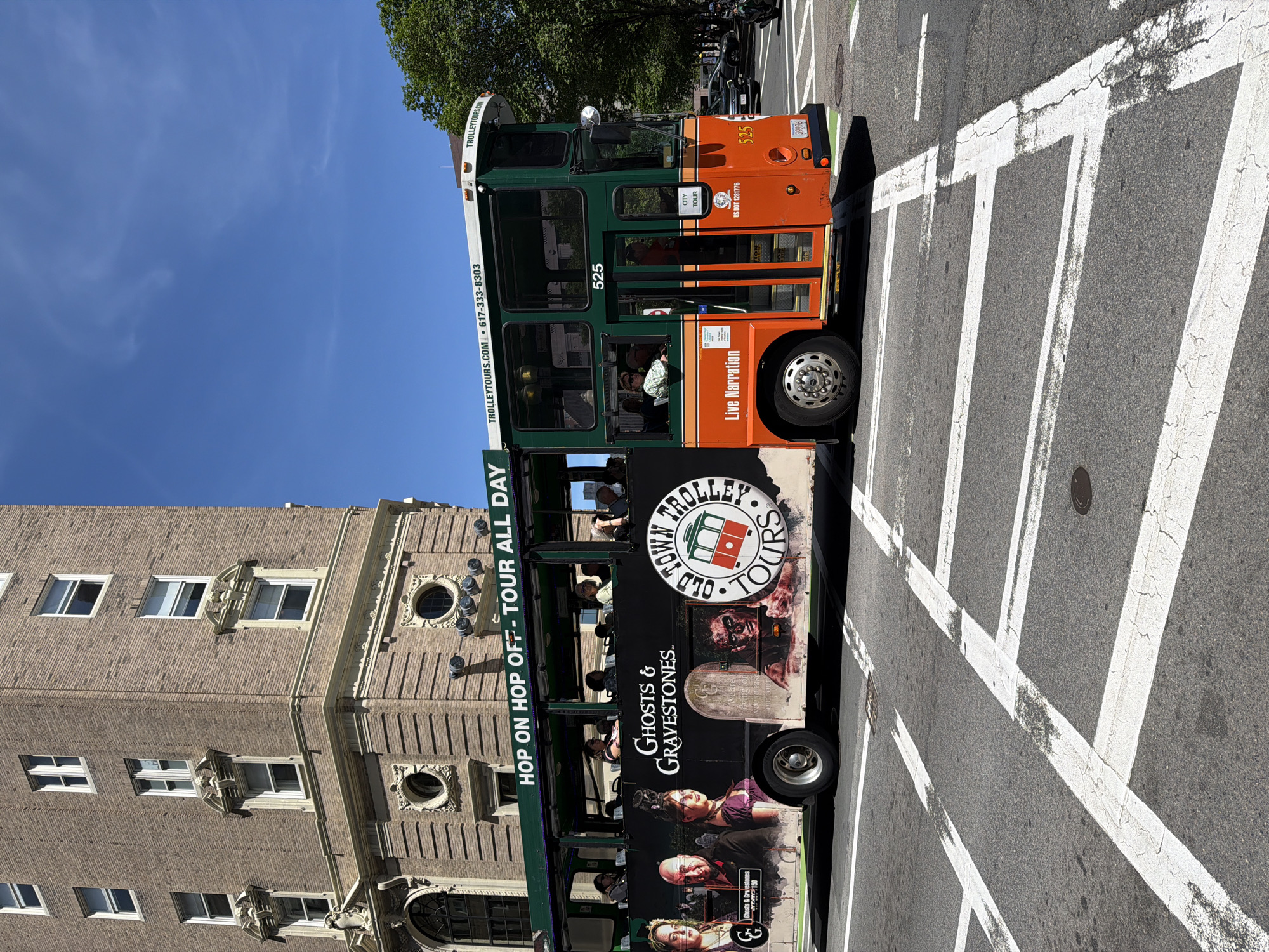 The image captures a green-and-orange Old Town Trolley Tours bus in Boston, Massachusetts, United States. The bus, numbered "525," is positioned on a road marked with white pedestrian crosswalk lines. The side of the bus features an advertisement for a "Ghosts & Gravestones" tour, complete with images of eerie characters and tombstones. The text "HOP ON HOP OFF-TOUR ALL DAY" is prominently displayed. The bus is occupied by passengers. A brick building with arched windows is visible in the background. The day appears sunny, with a clear blue sky. The trolley's tire is clearly visible.