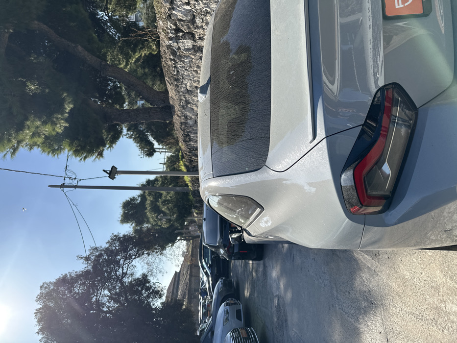 The image, taken in Metamorfosi, Greece, captures the rear passenger side of a light grey car, possibly a hatchback or compact SUV, parked outdoors on a bright, sunny day. The vehicle is covered in a noticeable layer of dust or pollen, particularly visible on its rear window and body panels. Its modern taillight features a dark housing with red and clear light elements. A line of other parked cars is partially visible behind the primary vehicle. In the background, there are several trees, including tall conifers and smaller deciduous trees, and a stone or rocky wall. Overhead, power lines run between utility poles against a clear blue sky. The angle of the sun casts long shadows, indicating it is daytime. On the upper right section of the main car's rear, a red rectangular object, possibly a sticker or part of a license plate, shows faint, unreadable white text. No specific activity is occurring.