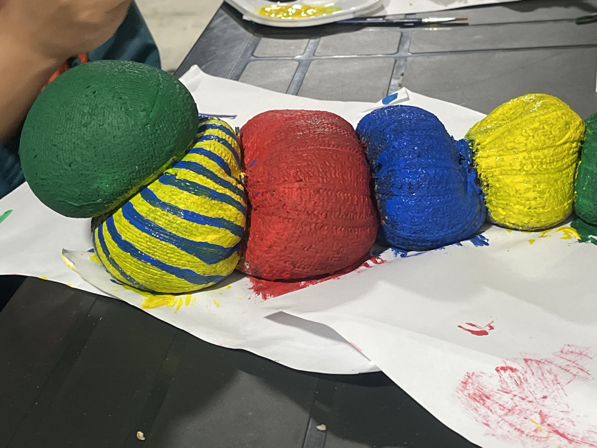 The image depicts a close-up, overhead view of several painted rocks arranged on a white paper surface, likely in an art or craft setting. The rocks are of various colors: green, yellow with blue stripes, red, and blue. To the far right, partially visible, is a yellow rock and possibly another green rock. The rocks have a textured appearance, suggesting they may be covered in yarn or twine before being painted, or the paint itself has created this texture. Splatters and smudges of paint, particularly red and yellow, are visible on the white paper and the dark surface below. In the upper background, a person's arm and shoulder, wearing a teal garment, are visible, suggesting someone is actively engaged in or observing the activity. A white plate with dollops of yellow paint and a paintbrush are also present in the background, further indicating an art project. The scene is set on a dark, possibly a table, surface with a patterned inlay. The lighting is diffuse, casting soft shadows. The image does not provide any information regarding the city of Surami, Georgia.
