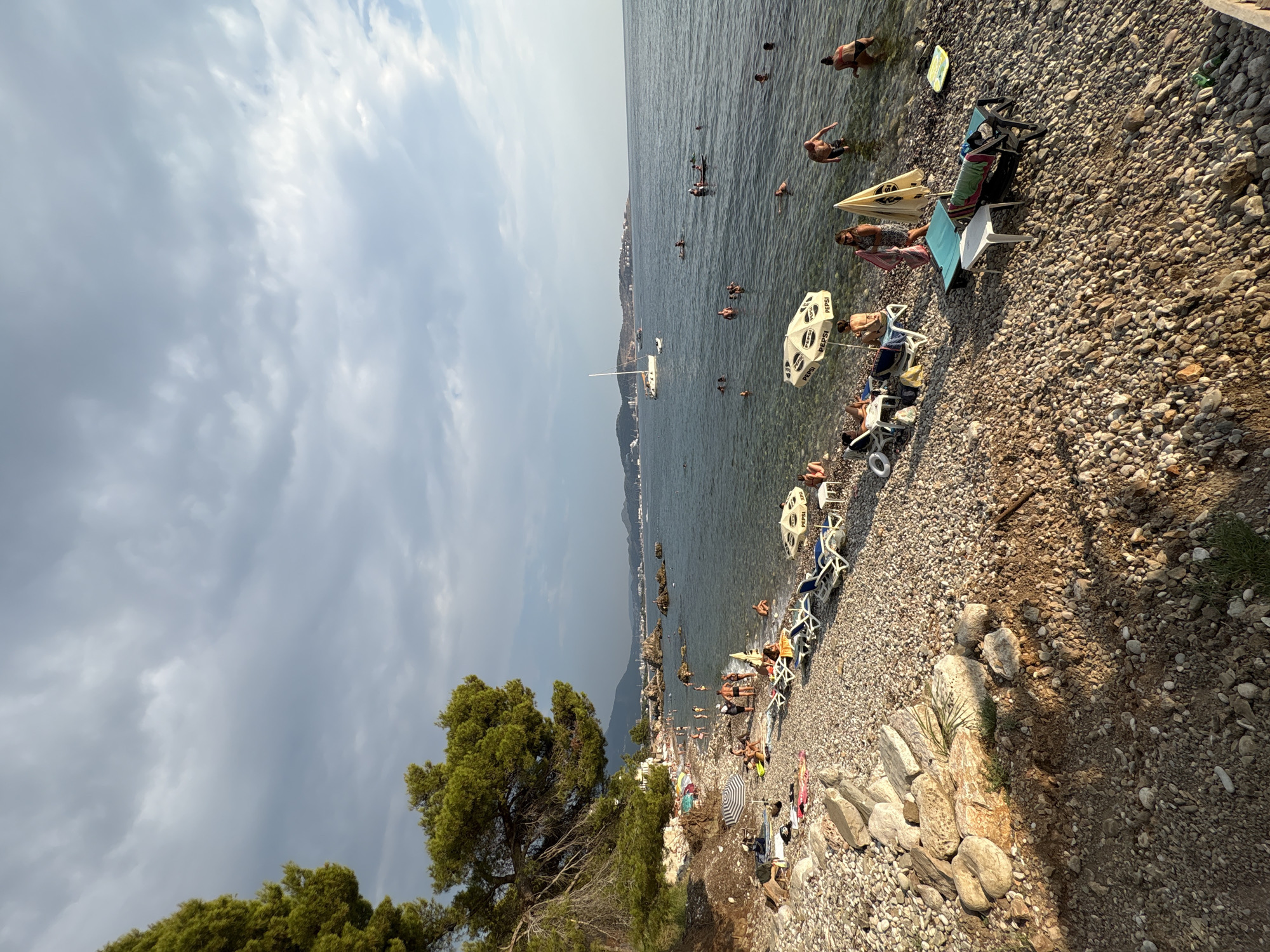 This is a vertical, eye-level, medium-shot, candid photograph taken on a pebbled beach in Bar, Montenegro, during daylight hours under a partially cloudy sky. The foreground is dominated by a section of the beach with many small, smooth pebbles and larger rocks. A large pine tree with green needles extends from the upper left corner, casting shadows.
In the midground, the beach continues with a scattering of lounge chairs, some occupied by people, and beach umbrellas. One umbrella is white with "PEPSI" branding. Several people are visible on the shore and in the shallow water, some wading, some swimming, and others interacting. Further out in the sea, there are more people swimming, and a white sailboat is visible in the distance. The water is clear and calm.
The background consists of a hazy coastline with hills and buildings, suggesting a coastal town or city. The sky above is a mix of gray and white clouds, with some patches of blue sky visible, indicating a dynamic weather condition. The overall atmosphere is that of a relaxed summer day at the beach.