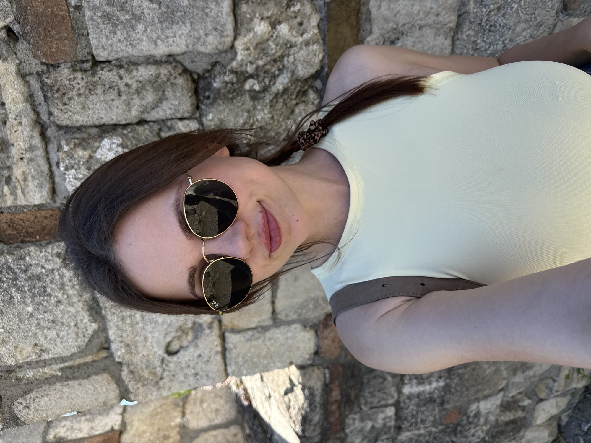A medium shot depicts a young woman posing for a selfie against a weathered stone wall. The woman is fair-skinned, with light-red lipstick and dark brown hair pulled back into a ponytail with a patterned scrunchie. She wears sunglasses with gold frames and a pale yellow shirt. A brown strap is visible across her body. The stone wall is made up of irregular, light-colored stones of varying sizes. The location is the Belgrade Fortress in Belgrade, Serbia.