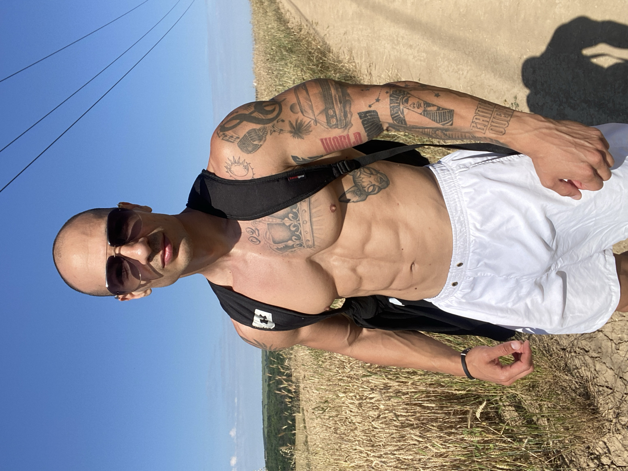 The image depicts a muscular, bald man with sunglasses and a black backpack standing in a rural setting. He is wearing white shorts and a black tank top, with numerous tattoos visible on his chest and arms. The man is standing on a dirt path next to a field of dry, golden wheat under a clear blue sky. Power lines are visible in the background. The scene appears to be a sunny day in the countryside, possibly on the outskirts of Pleven, Bulgaria, given the agricultural landscape.