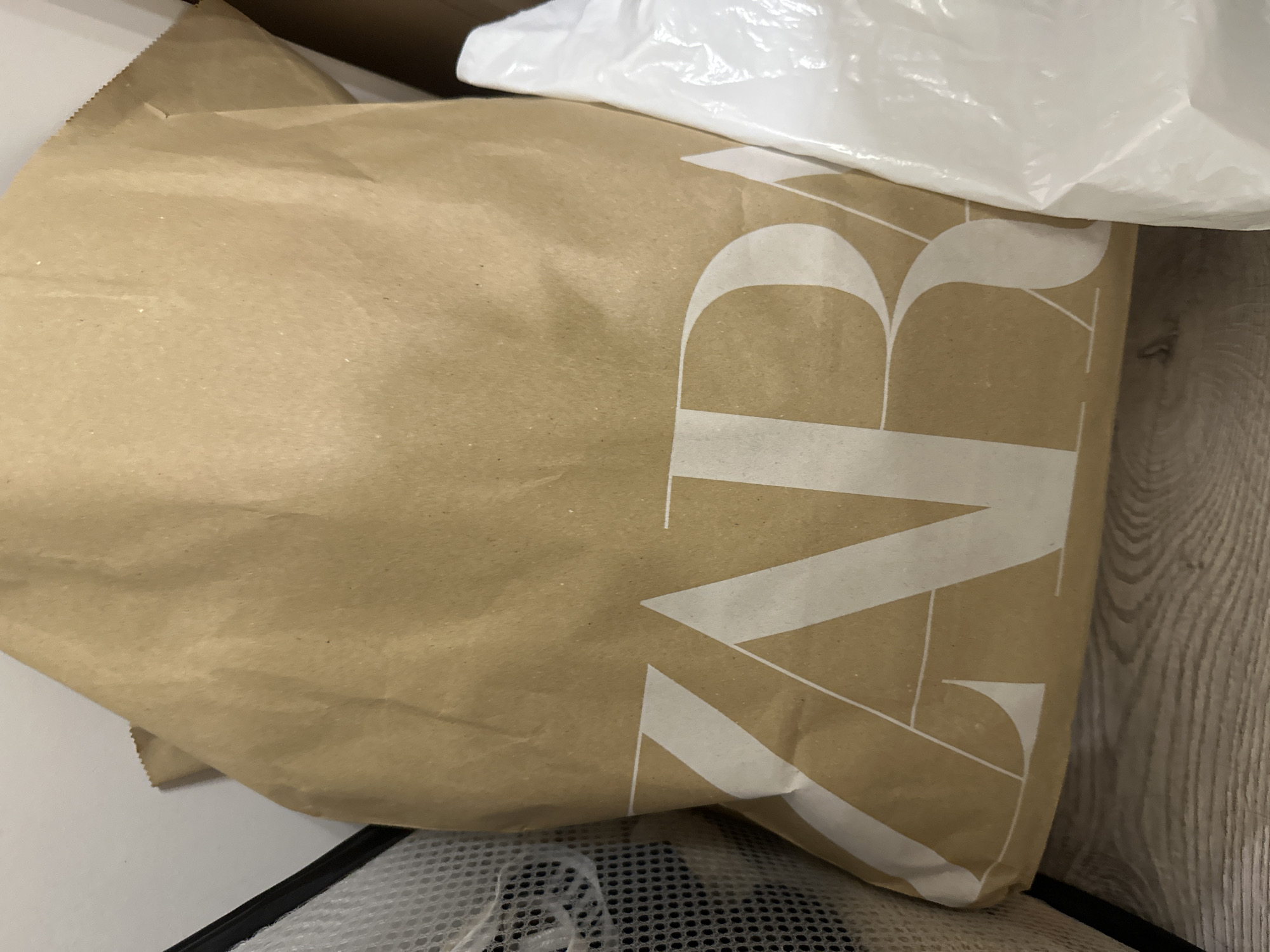 Location: Unknown, Unknown.
An indoor, static scene captured from a slightly elevated, frontal perspective. A large, light-brown kraft paper shopping bag, bearing the brand name "ZARA" printed in large white capital letters across its lower half, occupies the central foreground. The bag exhibits multiple wrinkles and creases, with fine, light-colored particulate matter scattered across its surface. Its upper edge shows a crimped, serrated detail. To the left of the ZARA bag, a portion of a black-rimmed, white mesh container, possibly a laundry hamper, is visible. To the right, a crumpled white plastic bag partially obscures the lower right section of the ZARA bag. The objects rest on a light-colored wooden floor with discernible grain patterns. A plain white or off-white wall forms the background.