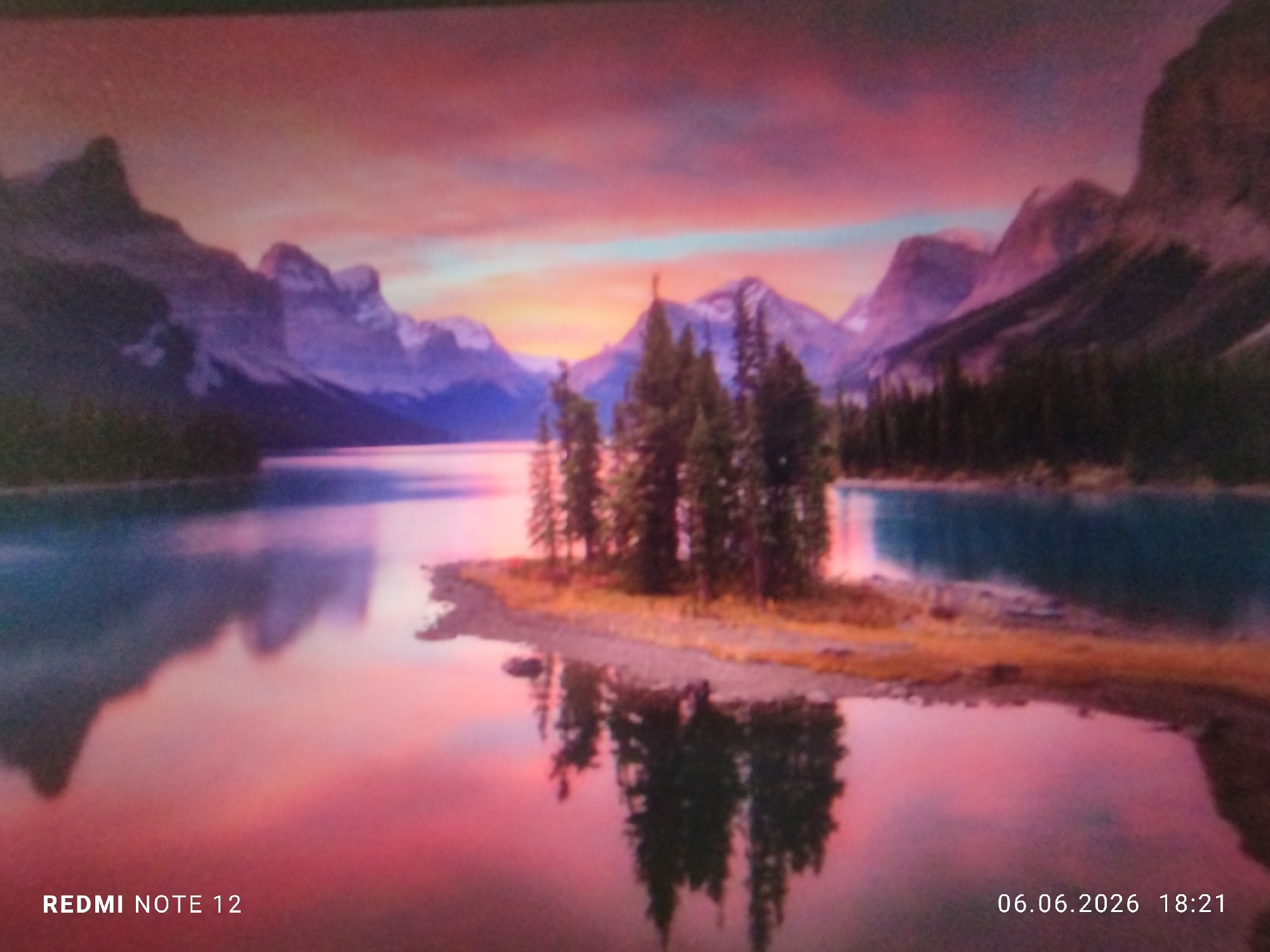 The image displays a panoramic landscape at sunset or sunrise, characterized by a vibrant interplay of colors. The sky transitions from deep red and pink at the zenith, fading into orange and light blue along the horizon. A prominent range of rugged mountains, appearing in shades of deep purple and dark blue with hints of snow on some peaks, forms the backdrop. In the foreground and mid-ground, a calm body of water, likely a lake, perfectly reflects the colorful sky and the mountain silhouettes, creating an almost symmetrical composition.
Centered within the water body, a small, irregularly shaped island or peninsula features a dense cluster of tall evergreen trees, predominantly dark green to black against the bright water and sky. The shoreline of this landmass exhibits earthy tones of brown and gold, suggesting dry grass or sandy terrain. Additional dark trees line the distant shores on both the left and right sides of the frame. The overall atmosphere is serene and natural. This scene is situated in the city of Burgas, Bulgaria. The photograph was captured on a Redmi Note 12 device on June 6, 2026, at 18:21. No individuals or active subjects are visible in the scene.