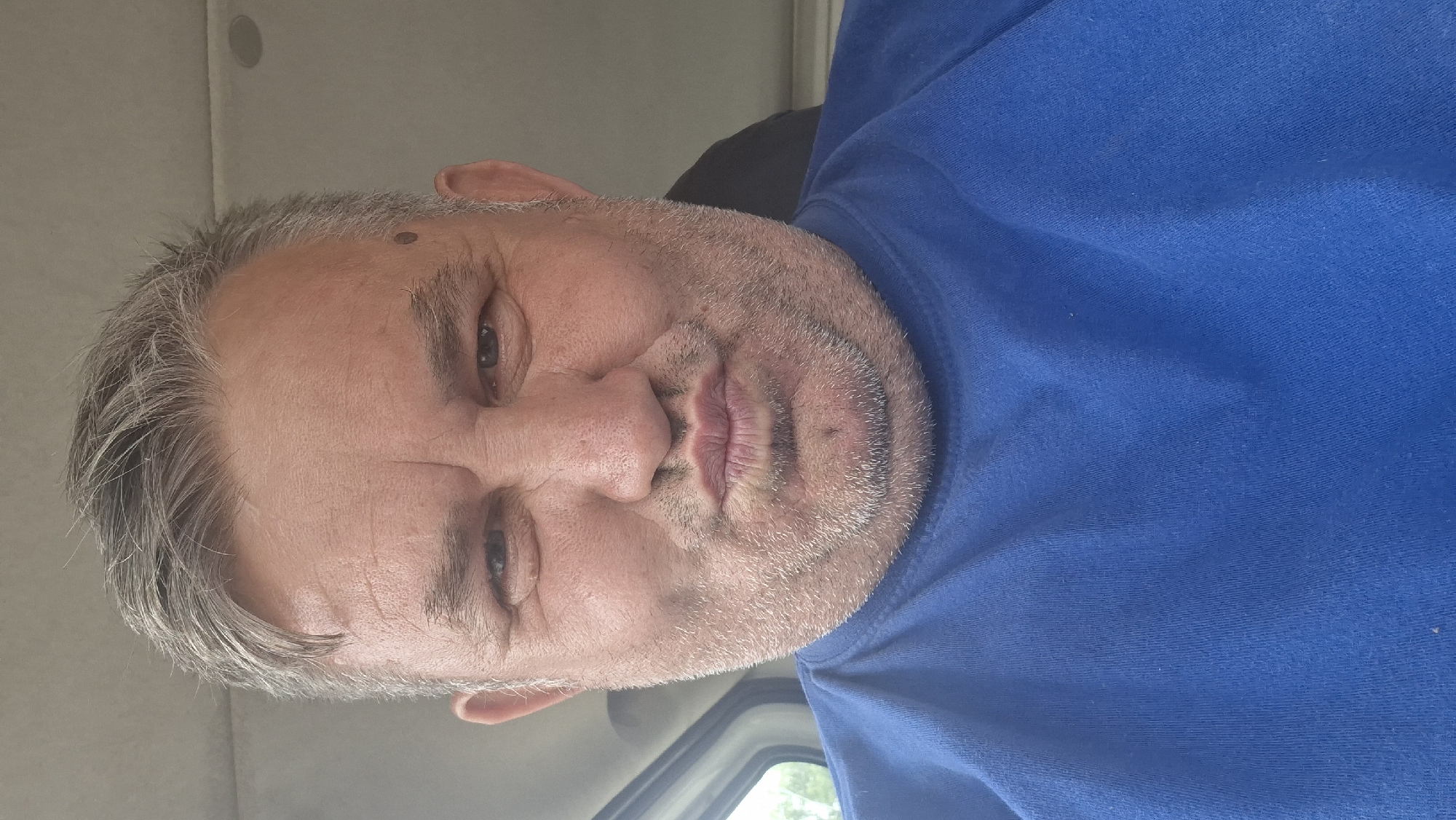 A medium shot selfie shows a man in his late 40s to early 50s with graying hair and a light beard. He is wearing a blue t-shirt and is making a duck face. The man is indoors, possibly in a vehicle, with a blurred background that suggests a window on the left and a car interior on the right. The image does not provide any specific clues to its location within Hîncești, Moldova.