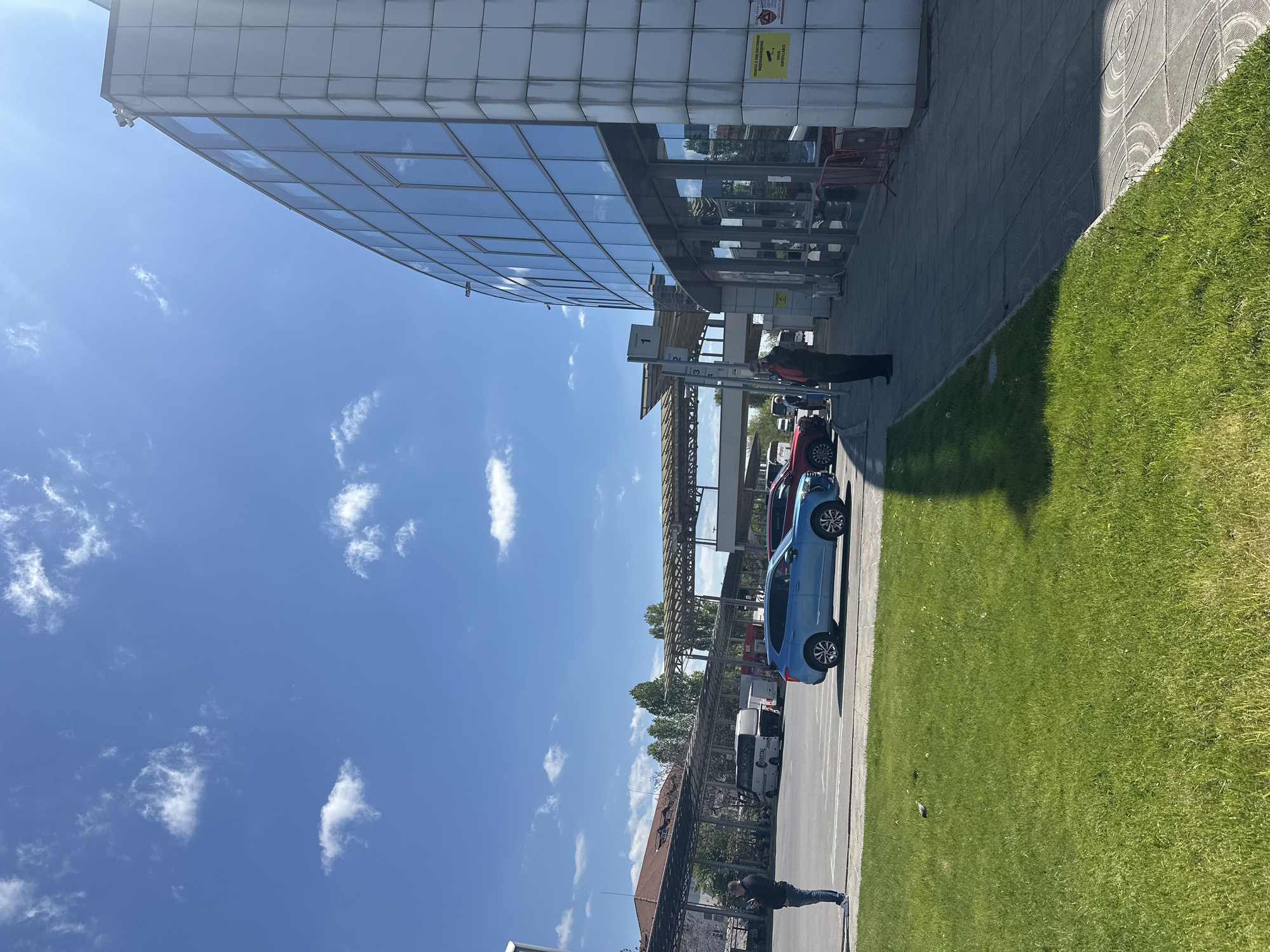 The image depicts an outdoor scene on a sunny day. A modern building with reflective glass windows is prominent on the left side. Adjacent to the building is a paved area or sidewalk, next to a grassy area.
Several cars are parked in a line, including a blue car and a red car. The scene also shows an open-air parking or waiting area covered with a metal structure.
Two people are visible: one near the building and another further down the parking area.
The sky is clear and blue with scattered clouds. Given the architectural style and environment, it is likely taken in a modern area of the city of Sofia, Bulgaria.