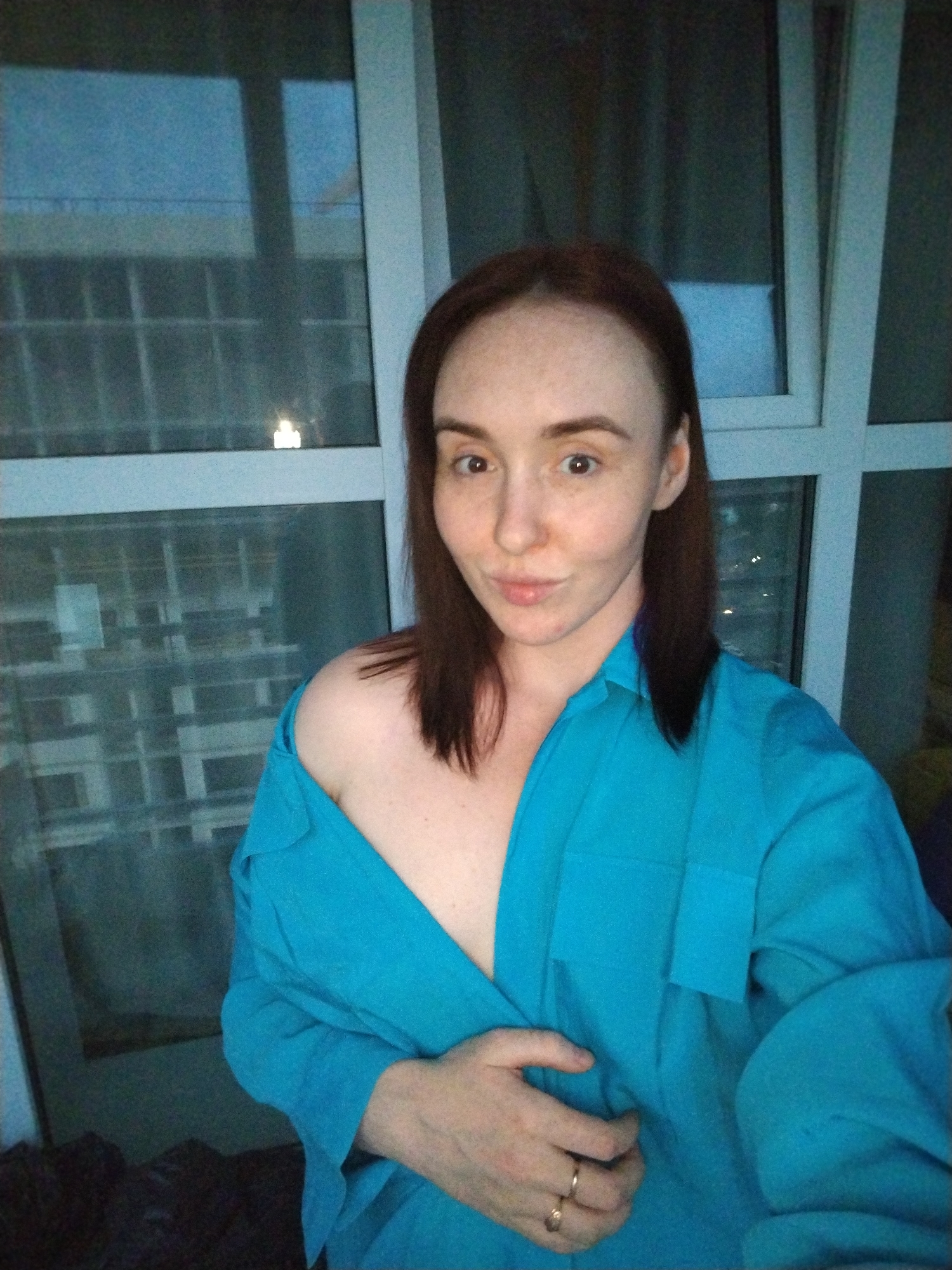A woman is posing indoors, possibly in an apartment, with a large window behind her. The window has a grid pattern and reflects the cityscape, potentially hinting at a location in a high-rise building. The reflection appears to show other buildings, with at least one light suggesting it might be nighttime or dusk.
The woman has fair skin, brown hair, and noticeable freckles. She is wearing a blue button-up shirt, which is partially unbuttoned, revealing one shoulder. She is holding the shirt closed with one hand and is wearing a ring on her finger. She is looking directly at the camera with a slightly puckered expression.
Without additional information about specific landmarks or architectural styles visible in the window reflection, it is not possible to definitively confirm the location as Izhevsk, Russia.