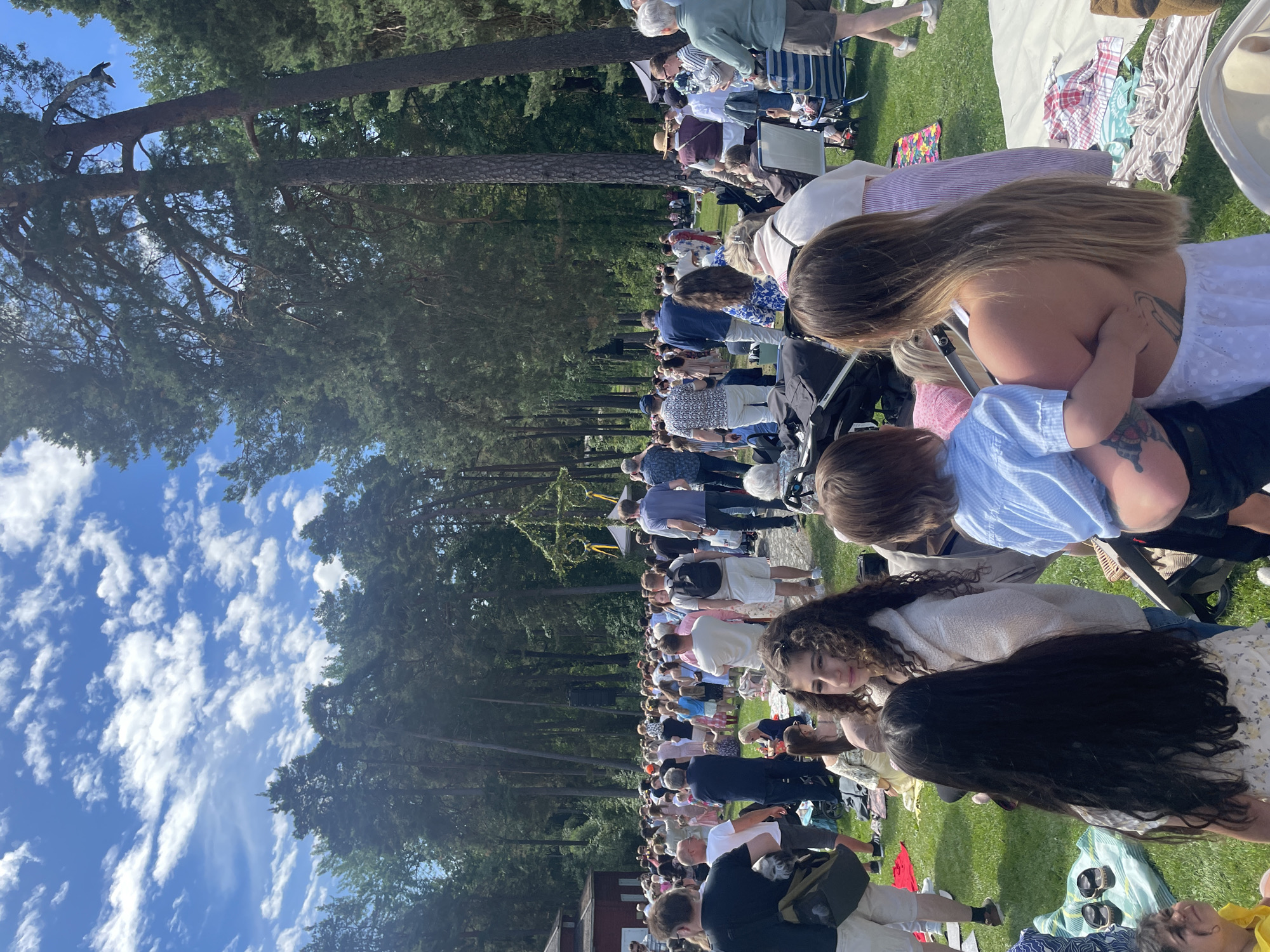 The image shows a large gathering of people in a park-like setting with tall trees and a blue sky with scattered clouds. The event appears to be a festive outdoor gathering, possibly a celebration or festival, given the presence of a maypole adorned with greenery and ribbons in the background. People are spread across a grassy area, with some sitting on blankets and others standing. Many are dressed in casual summer clothing. A red building is visible to the left, and tents and chairs are scattered throughout the scene, suggesting various activities or vendors. The overall atmosphere seems relaxed and social. The presence of the maypole strongly suggests this event is taking place during Midsommar, a major Swedish holiday. Given the maypole and the characteristic setting of a large park or open field with mature trees, this scene is likely taking place at Karlstad's Midsommar celebration, which traditionally occurs at Folkets Park.