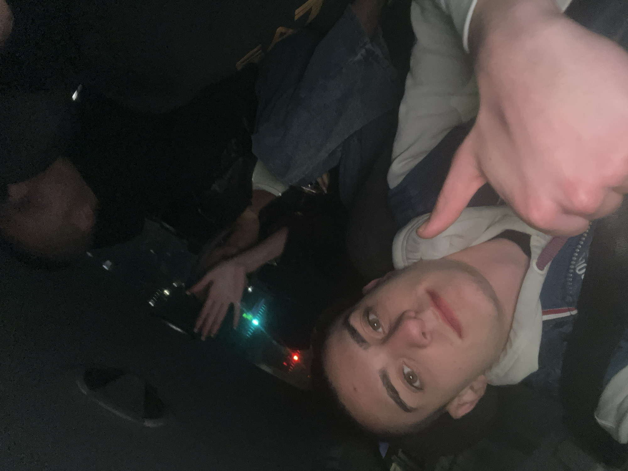 A photo taken inside a dark vehicle (possible limousine), containing at least three people. One man is visible in the foreground, angled towards the camera. The man has short brown hair, fair skin, and is wearing a light-colored hoodie with a blue jacket. His right hand is raised, pointing downward. Another person is visible in the background, partially obscured by the shadows. They are wearing jeans and a dark shirt. The third person is to the left, partially visible in the darkness. The interior of the vehicle appears dark, with hints of lighting equipment emitting green and red lights. The specific location in Burgas, Bulgaria is unknown.
