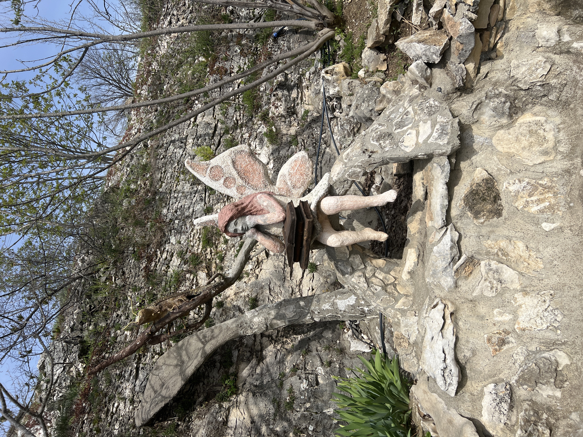 The image captures a fairy statue embedded in a rock wall or garden feature in Lukovit, Bulgaria. The statue is made of a rough, light-colored material, potentially concrete or plaster. The figure has wings decorated with pinkish-brown spots. The statue is partially obscured by a dark object, possibly a wooden structure. The surrounding environment is a rocky landscape with vegetation. Bare tree branches are visible in the upper left of the frame, with a blue sky visible through the branches. There is a structure below the fairy, with visible foliage and stones. The lighting is bright, suggesting a sunny day.