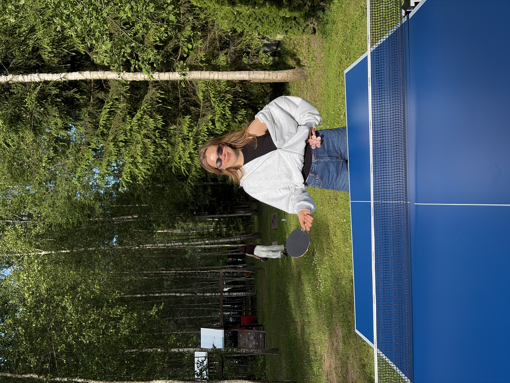 In the city of Zhdanovichsky Sel'skiy Sovet, Belarus, a young woman with blonde hair and sunglasses stands behind a blue ping pong table in a grassy yard, holding a ping pong paddle. She wears a grey hooded sweatshirt, partially unzipped, revealing a black top underneath, and blue denim shorts. To her left, in the background, a children's play structure is visible, and another person walks away from the camera. The yard is surrounded by tall trees, including birch and evergreen varieties, under a clear sky. The scene is bathed in the warm light of what appears to be late afternoon or early evening.