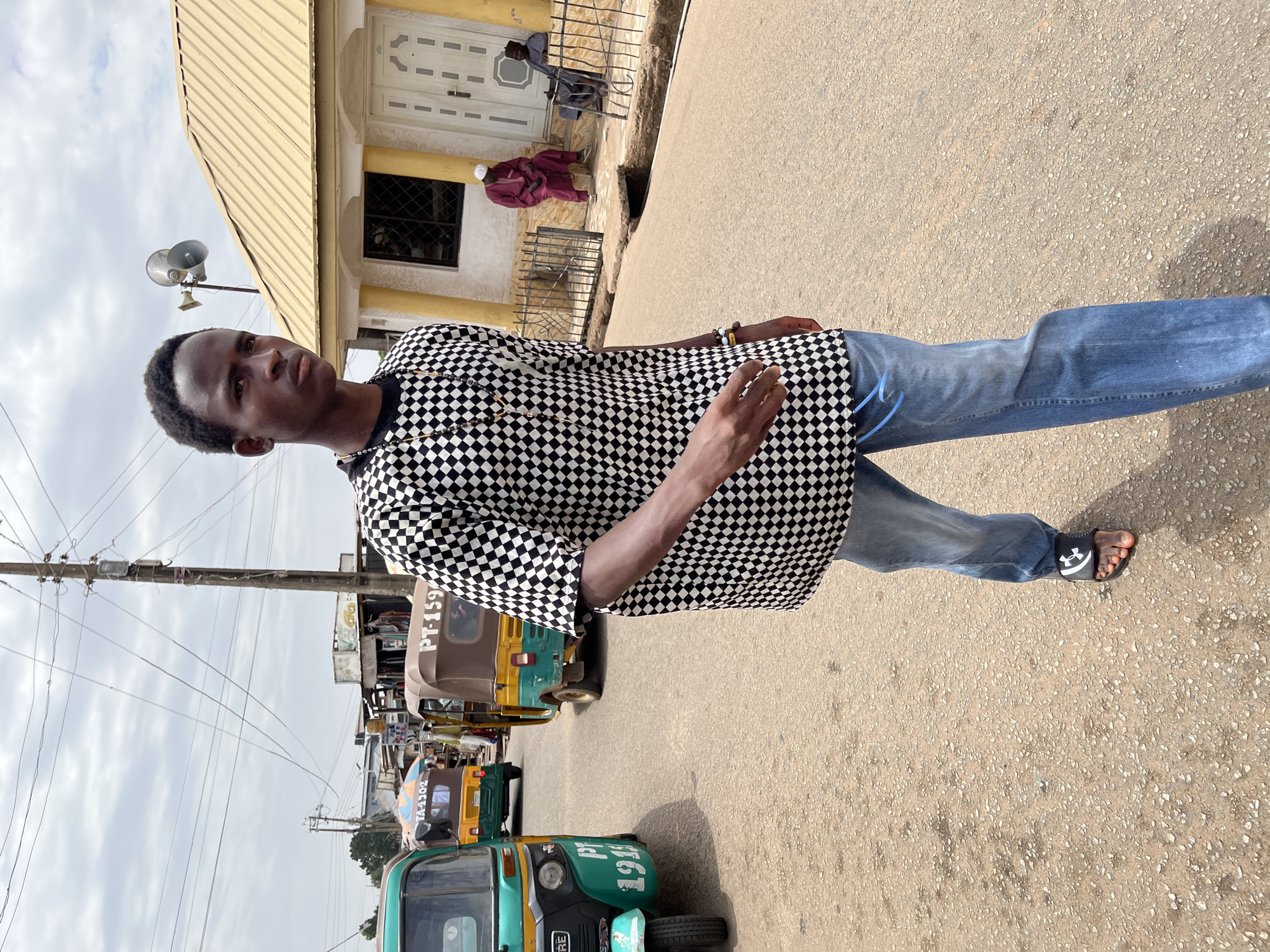 A man stands in the street in Gura Top, Nigeria. He wears a black and white checkered shirt, blue jeans, and Under Armour sandals. He faces the camera and looks directly at the viewer. Behind him are several auto-rickshaws. In the background, there is a building with a yellow roof and facade. Two men in traditional robes sit near the building's entrance. Overhead utility lines stretch across the sky. The sky is overcast. The road appears to be paved but weathered.