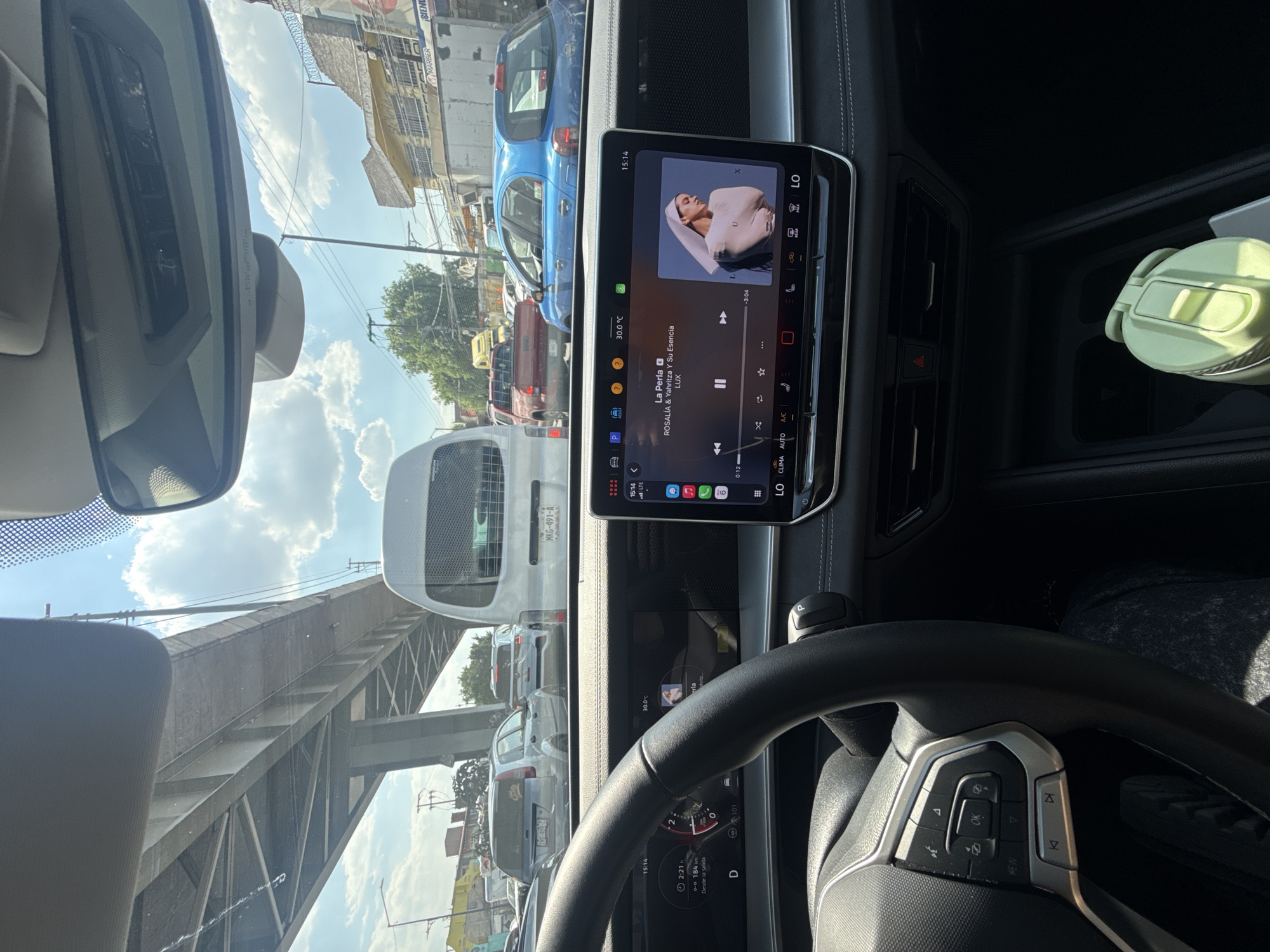 The image, taken from inside a car, shows a view of traffic in what appears to be a busy urban area. The car is stationary or moving slowly, indicated by the "D" on the dashboard and the slow speed of 0. The infotainment screen displays music by Rosalía and Yahritza Y Su Esencia playing on a music streaming service. Outside the car, traffic includes various vehicles, with a van and several cars visible. In the background, there's an elevated highway structure and buildings that suggest an urban setting.
The scene's location is in Mexico City, Mexico, as indicated by the license plate "MKG-491-A" and the presence of the Marber signage. The time on the car's display is 15:14 (3:14 PM).