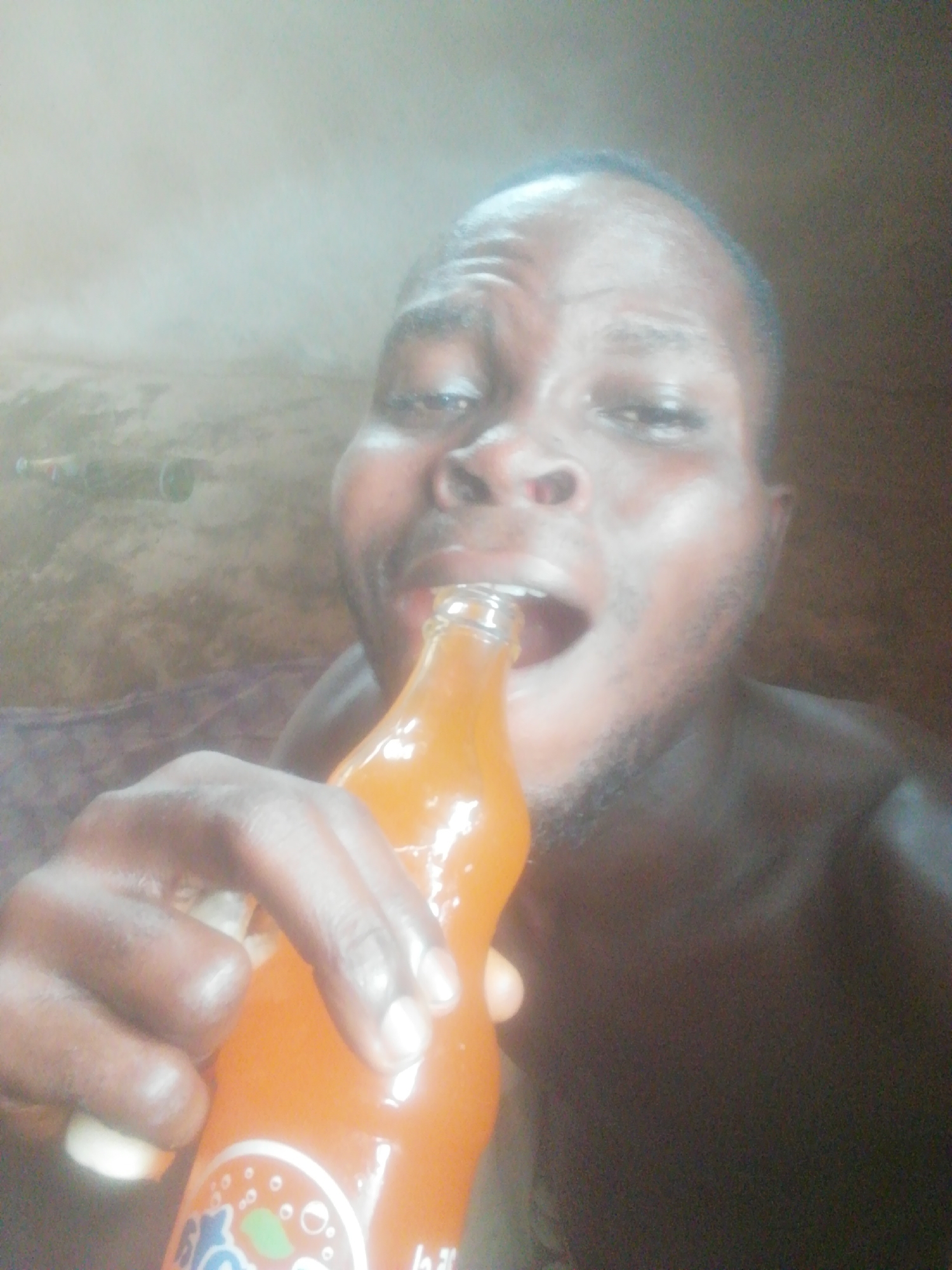 A man is drinking from an orange soda bottle, in a location of Unknown, Unknown. He is holding the bottle with his left hand and tilting it up to his mouth. The man has dark hair and a beard. The environment appears to be a room, with a blurry background.