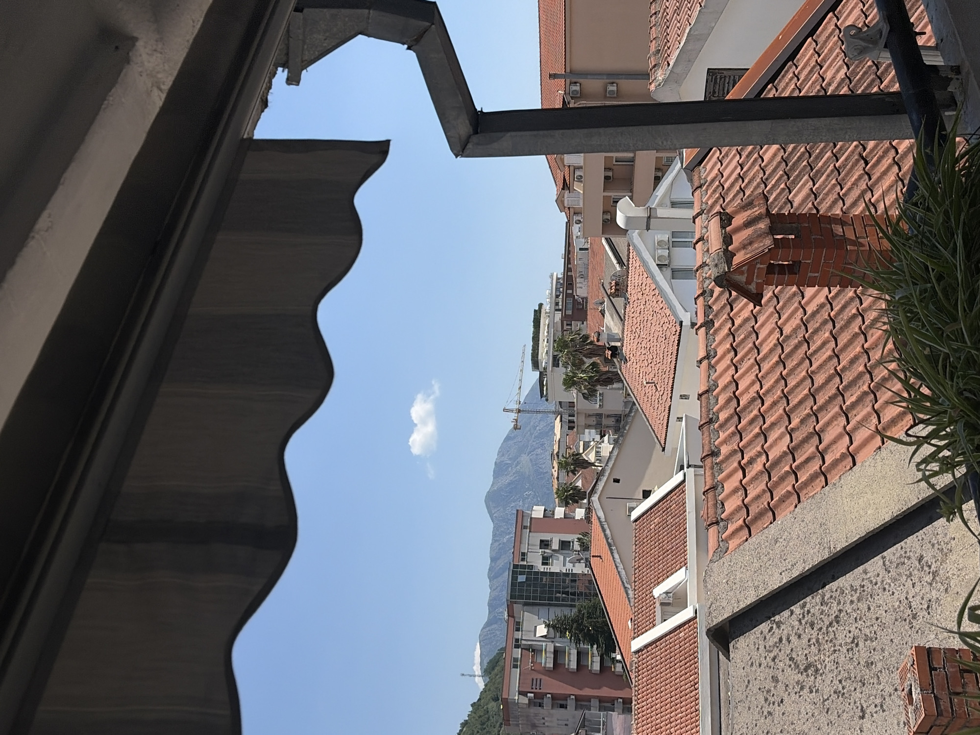 The image shows a view of the city of Budva, Montenegro, from a balcony or window, looking out over a neighborhood of buildings with red-tiled roofs. In the foreground, a dark awning with a scalloped edge partially obscures the sky. A dark metal gutter system is visible on the right side of the frame. Beyond the foreground, a cluster of residential buildings with red tile roofs dominate the scene. Some buildings are taller, multi-story structures, while others appear to be smaller houses or villas. A brick chimney with a decorative top is prominent on one of the roofs. In the midground, larger buildings with balconies and windows are visible, and a construction crane is present in the distance, suggesting ongoing development. The background features a range of mountains under a clear blue sky with a few wispy clouds. The overall impression is a sunny day in a coastal European city.