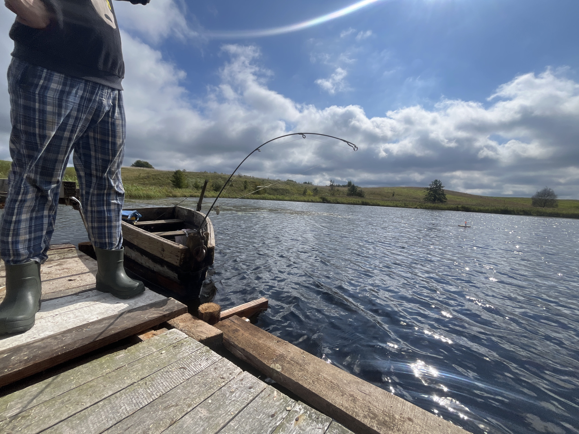 The image depicts a person standing on a wooden dock, fishing in a lake under a partly cloudy sky. The person is wearing plaid pajamas and green rubber boots. To the left of the person, a wooden fishing boat is tied to the dock. A fishing rod is bent, suggesting it is in use. The lake is calm with ripples on the surface, reflecting the sunlight. In the background, rolling green hills and scattered trees are visible. A fishing float is visible in the distance on the water. The scene appears to be in a rural outdoor setting, likely in the Дунілавіцкі сельскі Савет, Belarus.