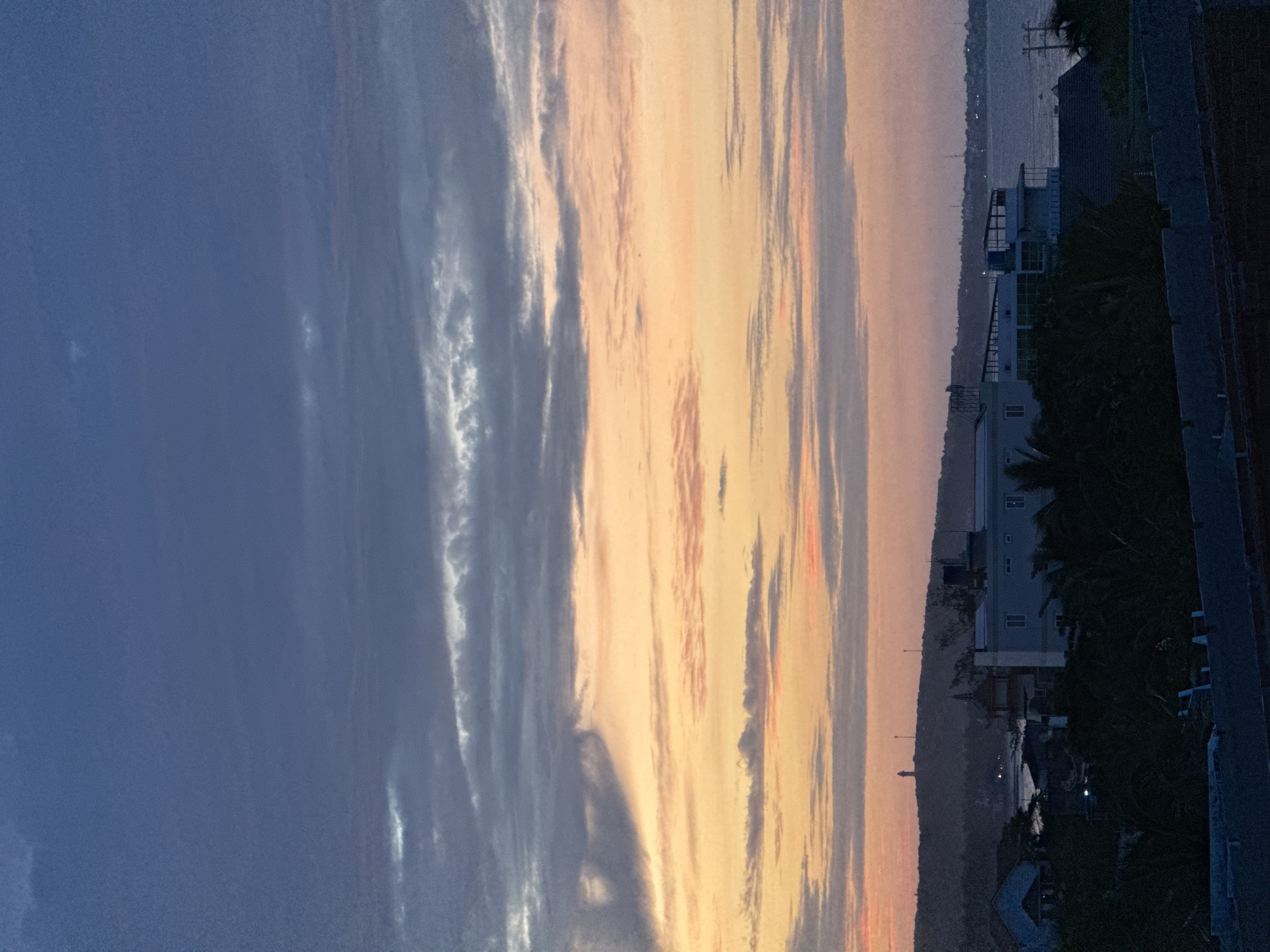 The image shows a twilight sky over Mũi Né, Vietnam. The sky transitions from dark blue at the top to hues of orange and pink closer to the horizon, with scattered clouds. In the foreground, silhouetted palm trees and rooftops are visible. Beyond the vegetation, a cluster of buildings, including a multi-story white structure with balconies, is situated along the coast. In the distance, a hill is topped by a lighthouse and other structures, with the sea stretching out to the horizon. The scene depicts a coastal town at dusk.