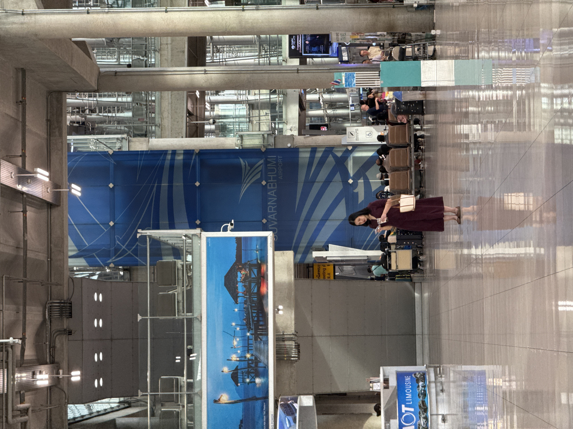 The image depicts the interior of a modern airport terminal, likely Suvarnabhumi Airport, located in the Nong Prue Subdistrict of Thailand. A woman in a burgundy dress stands in the foreground, looking at her phone. The airport features a large blue graphic on a wall, an advertisement for "HOT LIMOUSINE" on the left, and seating areas for passengers in the background. The floor is highly polished, reflecting the overhead lighting and the surrounding architecture. Several other individuals are visible, some seated and some walking, with luggage carts and trash receptacles present. The overall environment suggests a busy, functional travel hub.