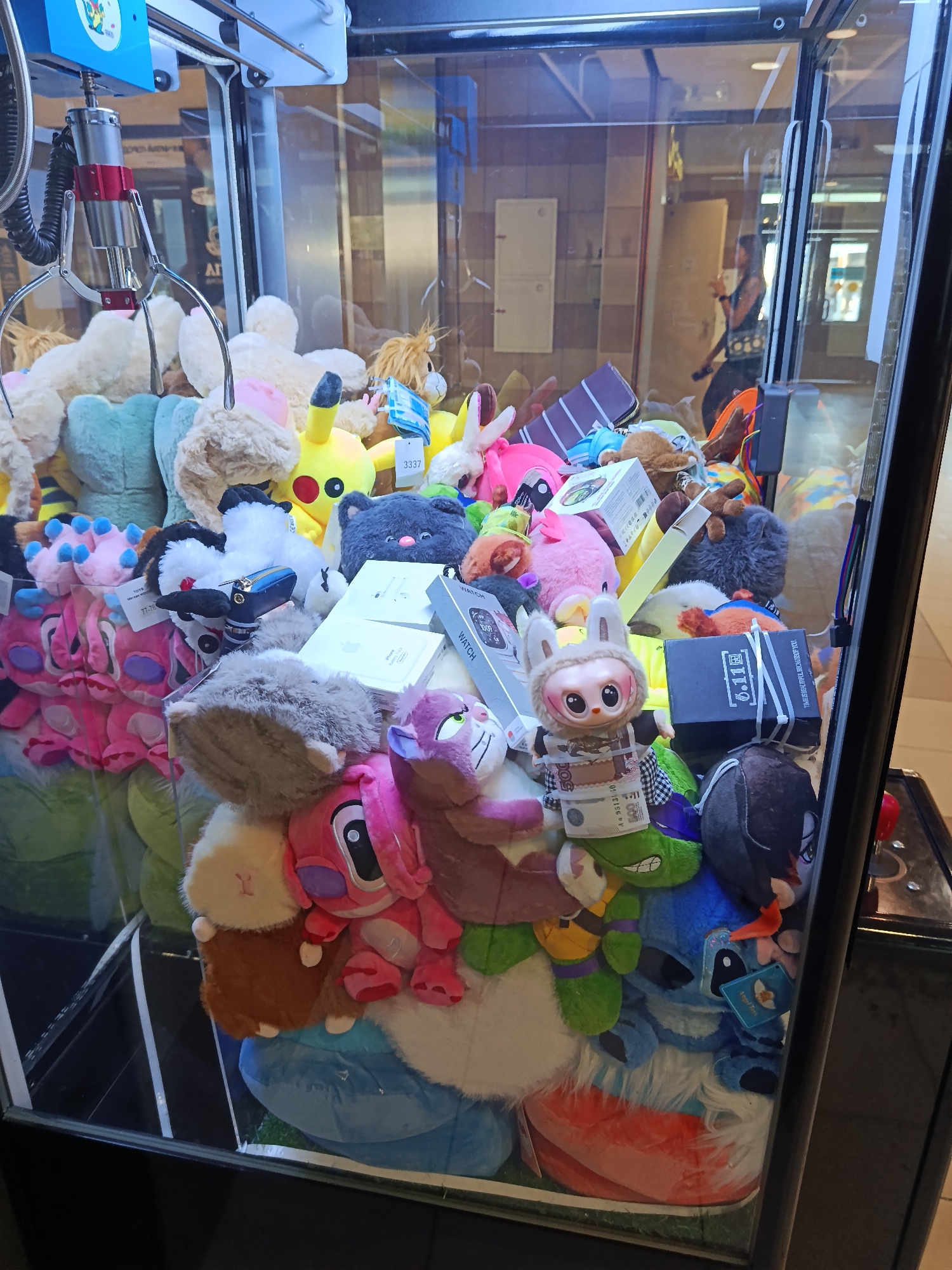 The image shows a claw machine filled with various stuffed toys and some boxes. The toys include characters like Pikachu and Stitch, as well as generic plush animals. Several boxes are visible, one labeled "WATCH" and another with Russian text and currency. The claw mechanism is visible at the top, with a metal claw suspended above the toys. The background appears to be an indoor public space, possibly a mall, with tiled walls and distant figures of people. The lighting is bright, suggesting an indoor environment. The overall scene depicts a typical claw machine arcade setup. The image is not specific to Moscow, Russia, and does not contain any identifiable landmarks or context that would place it in that city.