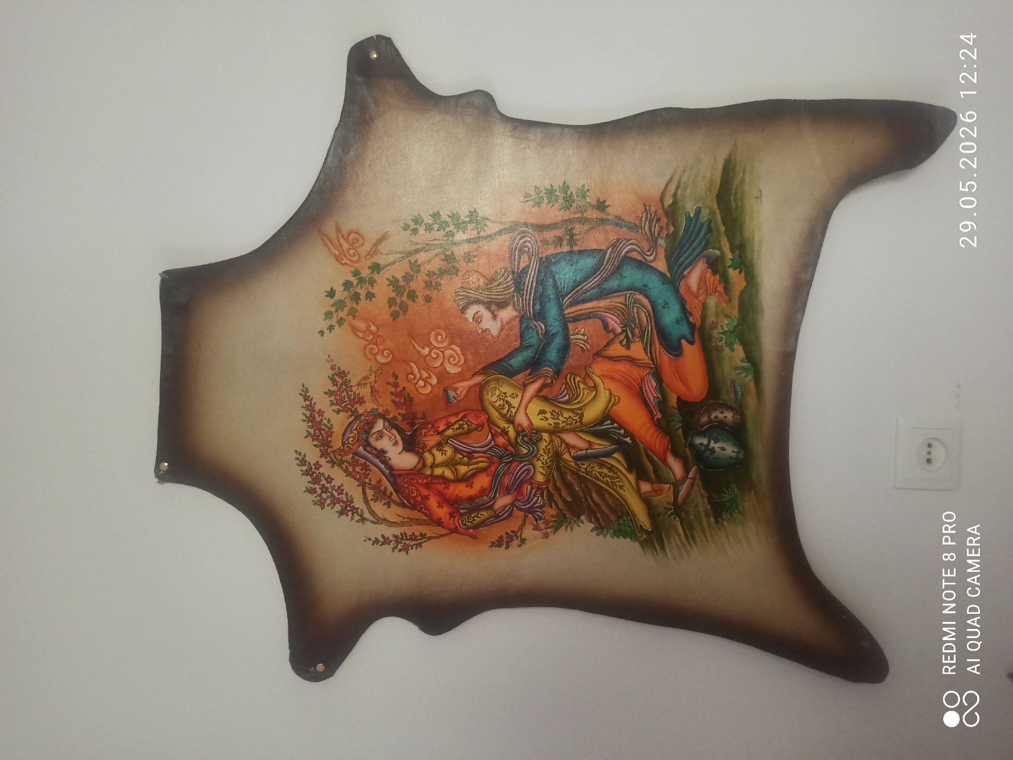 A piece of decorated leather hangs on a white wall. The leather is roughly cut in an irregular shape, with a dark brown burnt-looking edge. Painted on the leather is a scene in a traditional Persian miniature style, featuring two figures. A woman reclines in a red and gold gown, surrounded by flowers. A man leans over her, wearing a blue jacket and orange pants. They are under the branches of a tree in a landscape setting. The scene is detailed and colorful. The wall is white. A date and time stamp, "29.05.2026 12:24", are visible in the upper right corner. A switch plate is also visible on the wall. The location is Unknown, Unknown.