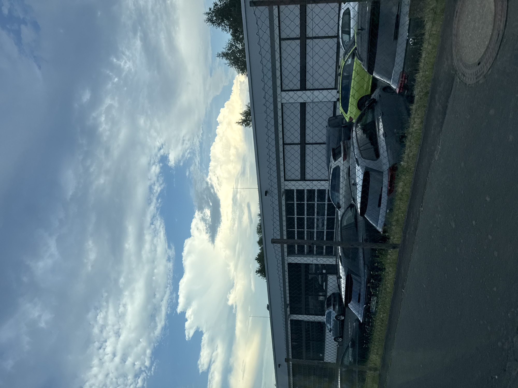 The image depicts a car dealership in Rechtenbach, Germany, under a cloudy sky. A row of cars, including a bright green station wagon, a dark gray SUV, and a silver car, are parked in front of a modern building with large glass garage doors. The building appears to be a commercial establishment, possibly a car repair shop or showroom. A chain-link fence separates the parking lot from a paved area in the foreground, which shows a manhole cover. Trees line the horizon behind the building, suggesting a semi-rural or suburban setting.