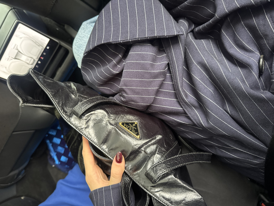 The scene is set in the rear passenger compartment of a vehicle in Unknown, Unknown. A person's right hand, with dark red manicured nails, holds a black, textured or patent leather Prada handbag, identifiable by its prominent gold triangular "Prada Milano Dal 1913" emblem. The bag is positioned over a navy blue garment with white pinstripes, likely a jacket or blazer, which is draped across a dark seat. A solid blue fabric is partially visible to the left. In the background, a digital screen on the back of the front seat displays graphic icons consistent with car seat controls, indicating a modern vehicle interior.