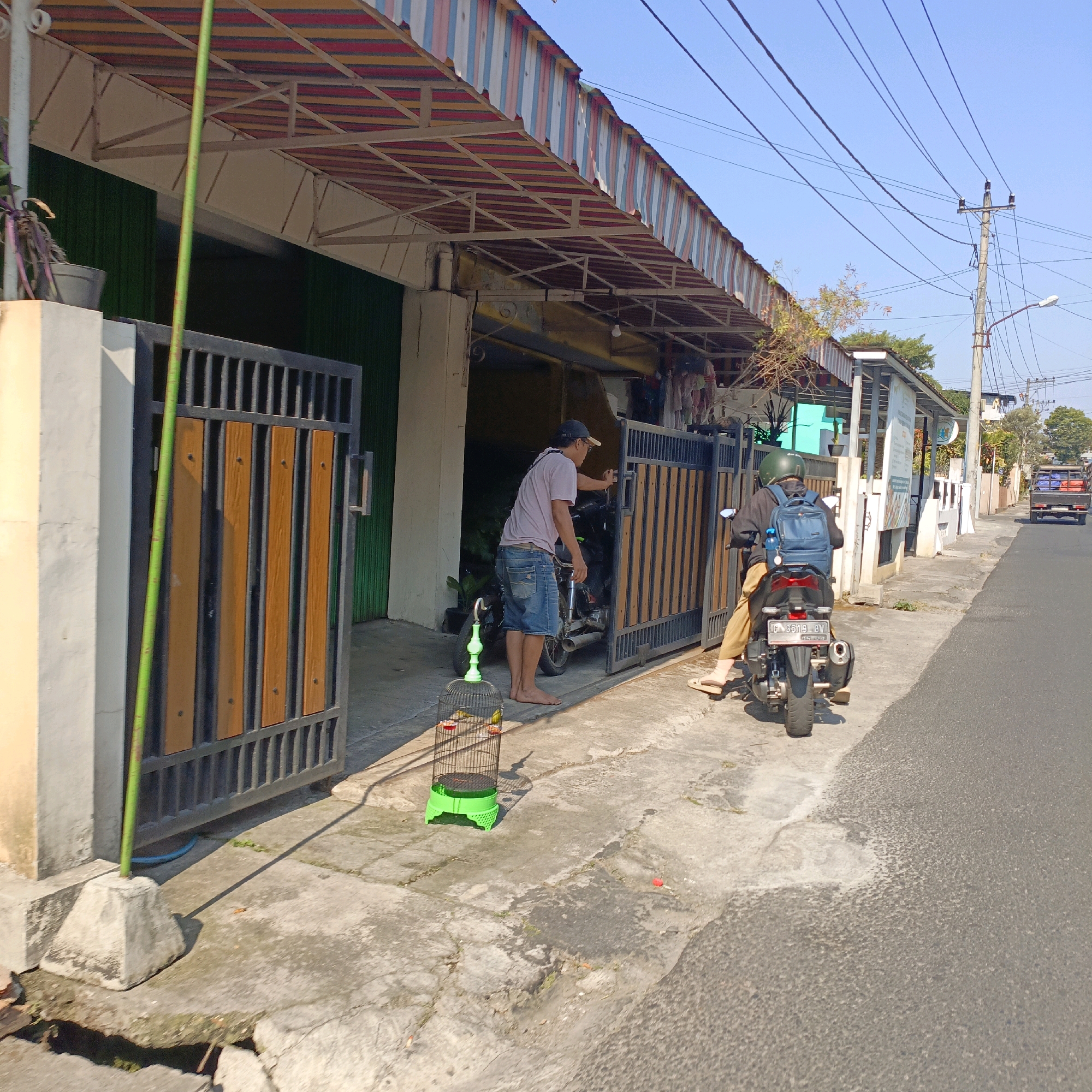 The image depicts a street scene in Salatiga, Indonesia, on a sunny day. In the foreground, a green bird cage with two birds inside sits on a concrete sidewalk. A man wearing a light purple t-shirt and denim shorts stands barefoot, facing away from the camera, near a closed metal gate with wooden slats. Behind him, a motorcycle is parked. To the right, another person, partially visible, is sitting on a motorcycle, wearing a black jacket, helmet, and tan pants. This motorcycle has a license plate that reads "G 3519 BV". The background shows buildings with striped awnings, a street with asphalt, and a utility pole with overhead wires. A truck with a load of goods is visible further down the street.