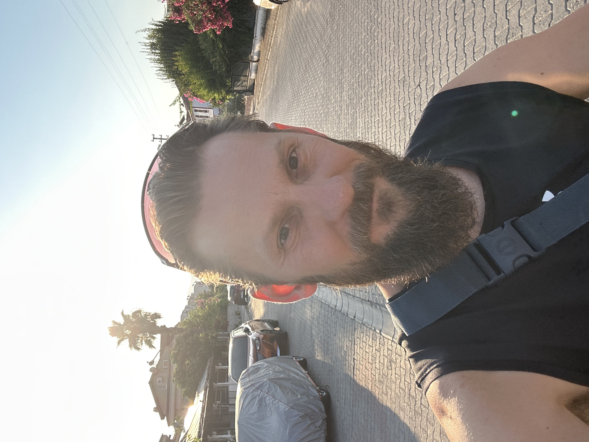 A man with a beard and sunglasses on his head takes a selfie in Fethiye, Turkey. He is wearing a black tank top and a grey strap is visible across his chest. The background shows a cobblestone street lined with parked cars and houses. A palm tree is visible in the distance. The sun is setting, casting a warm glow on the scene.