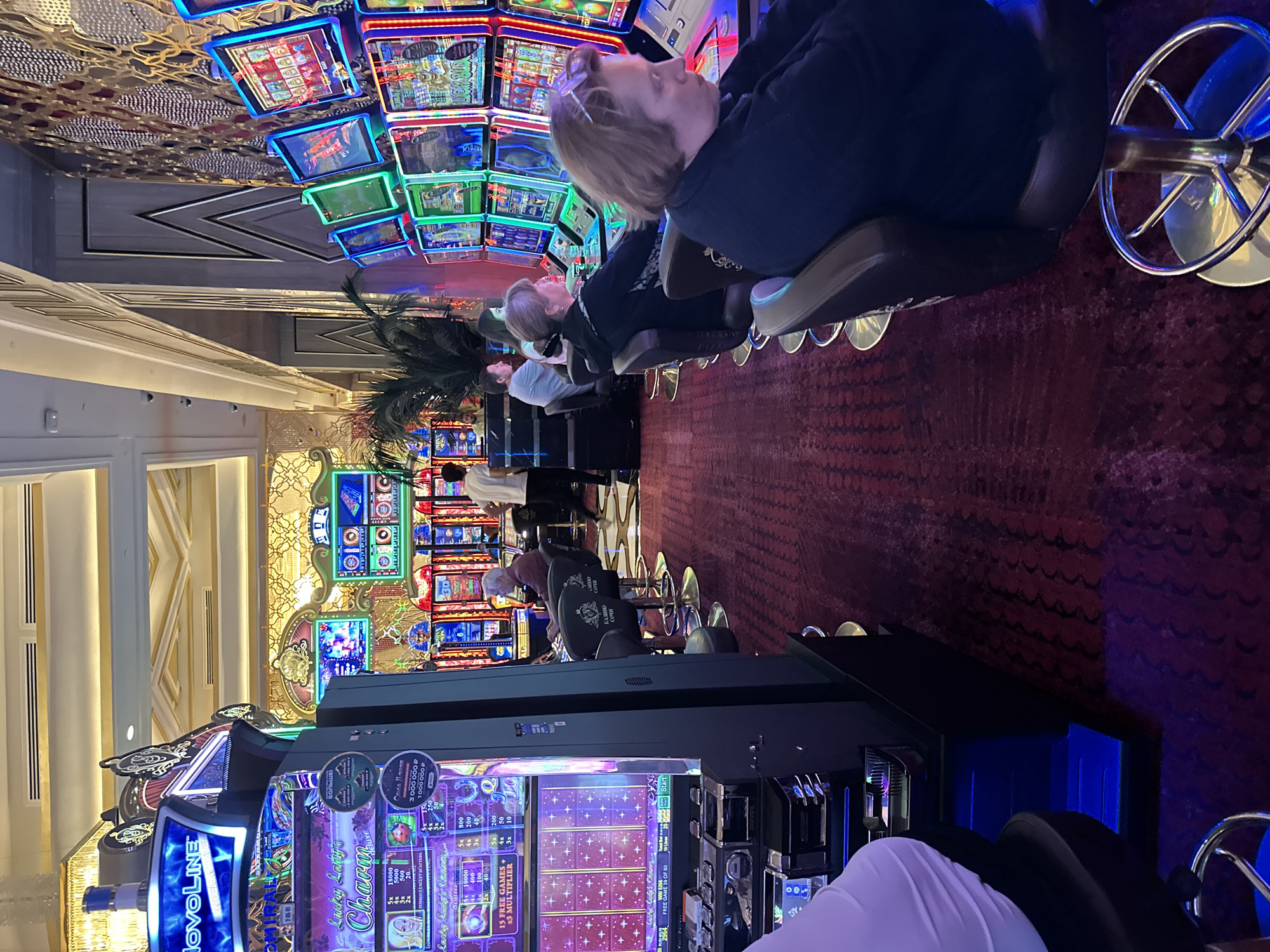 The image shows an interior scene in a casino located in Estosadok, Russia. The foreground features a woman seated at a slot machine, her hands on the controls. To her left, another woman is also seated at a slot machine, facing slightly away from the camera. Behind them, several other slot machines are visible, with people seated at some of them. The slot machines are brightly lit with colorful displays. In the background, more slot machines and a decorative wall are visible. The environment appears to be a well-lit, modern casino with a plush red carpet. The lighting is a mix of ambient casino lighting and the glow from the slot machines. The overall impression is one of activity and entertainment within a casino setting.
